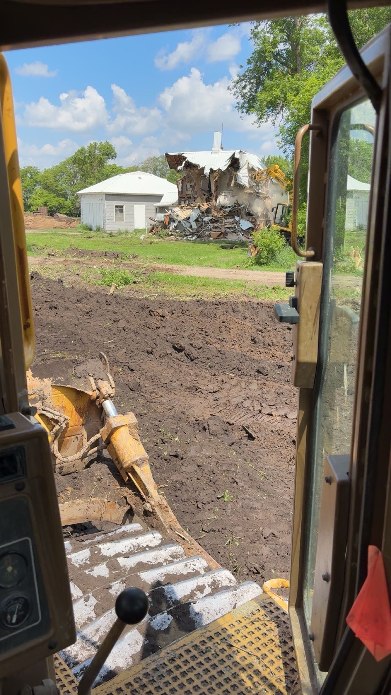 View from a bulldozer cab with a demolished building in the distance and a muddy landscape.