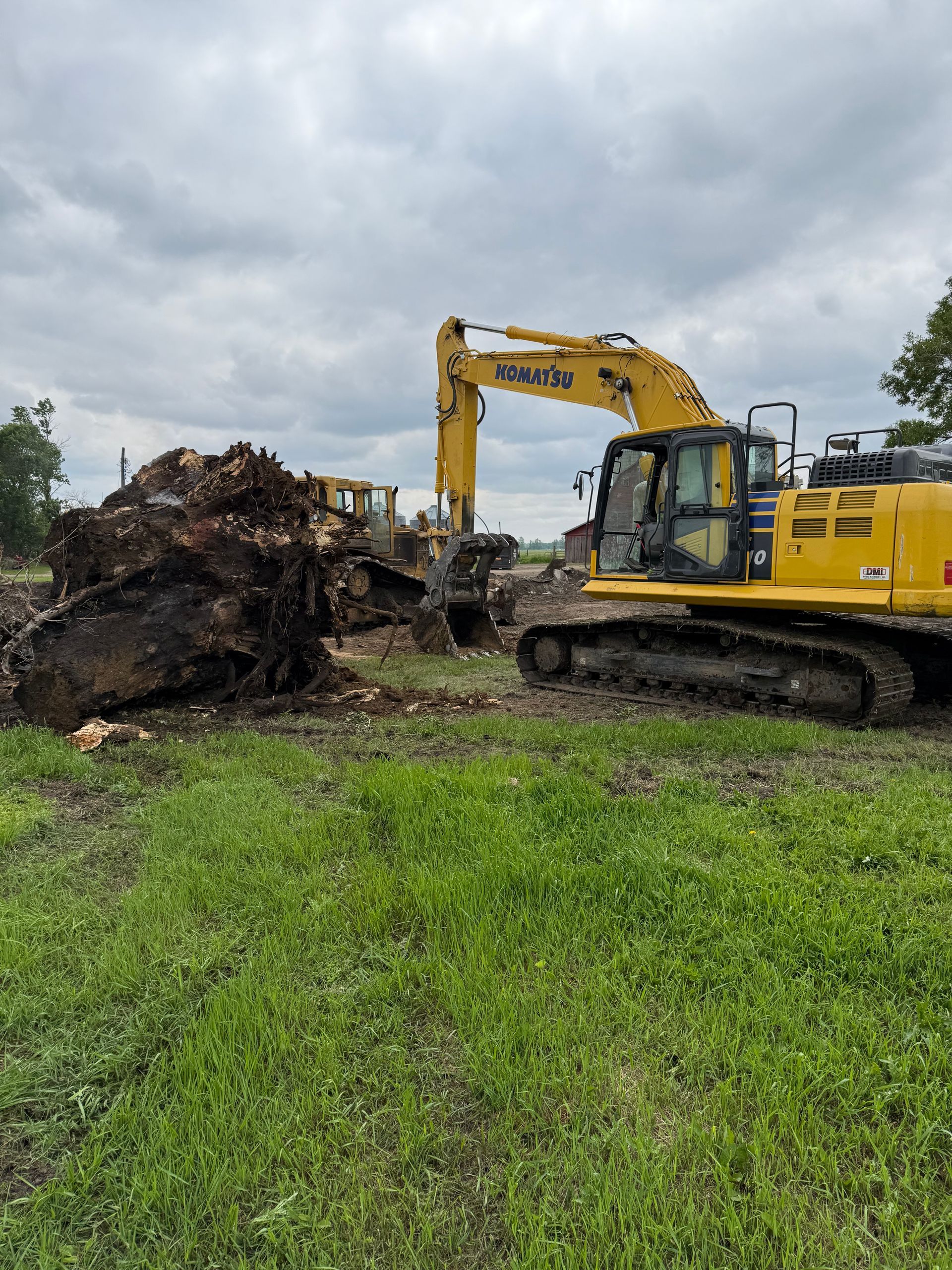 Yellow excavator near a large pile of tree roots on a grassy field, under a cloudy sky.