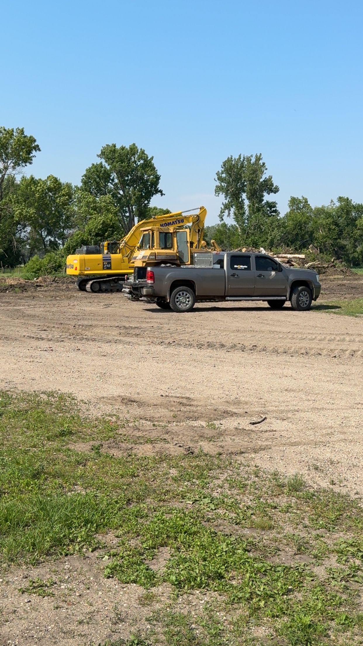 A yellow excavator and a gray pickup truck on a dirt road, trees in the background under a blue sky.