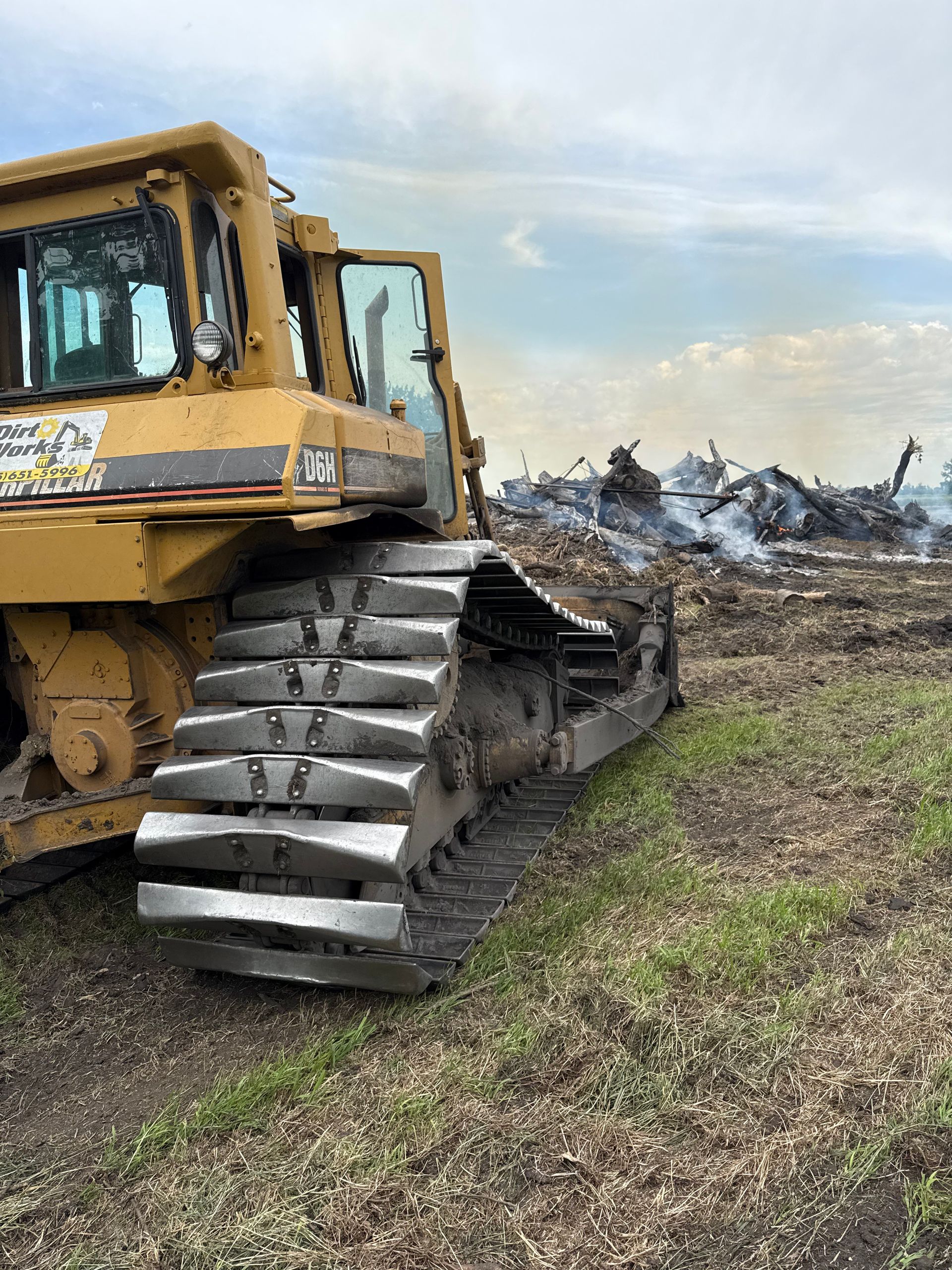 Yellow bulldozer on muddy ground, tracks in foreground. Burning debris in the background, cloudy sky.