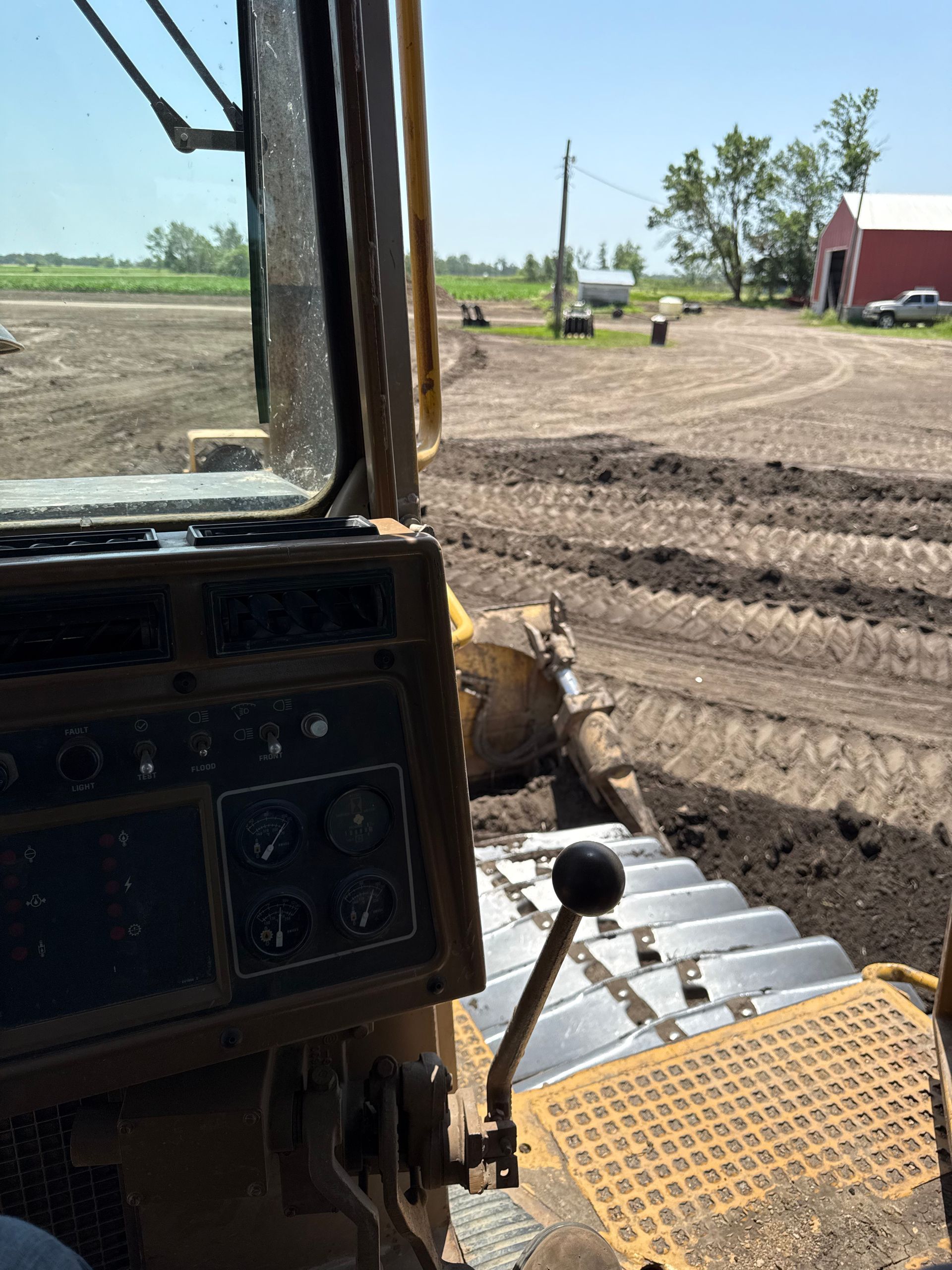 View from inside a bulldozer cab, grading soil in a field. Brown tracks, dirt, and control panel visible. Red barn in the distance.