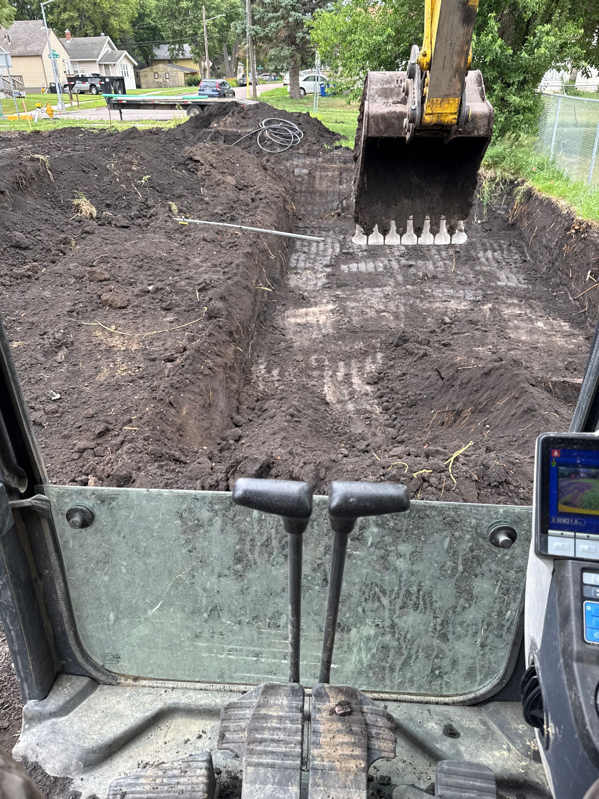 An excavator digs in a dirt area with the operator's view, a screen, and levers visible.