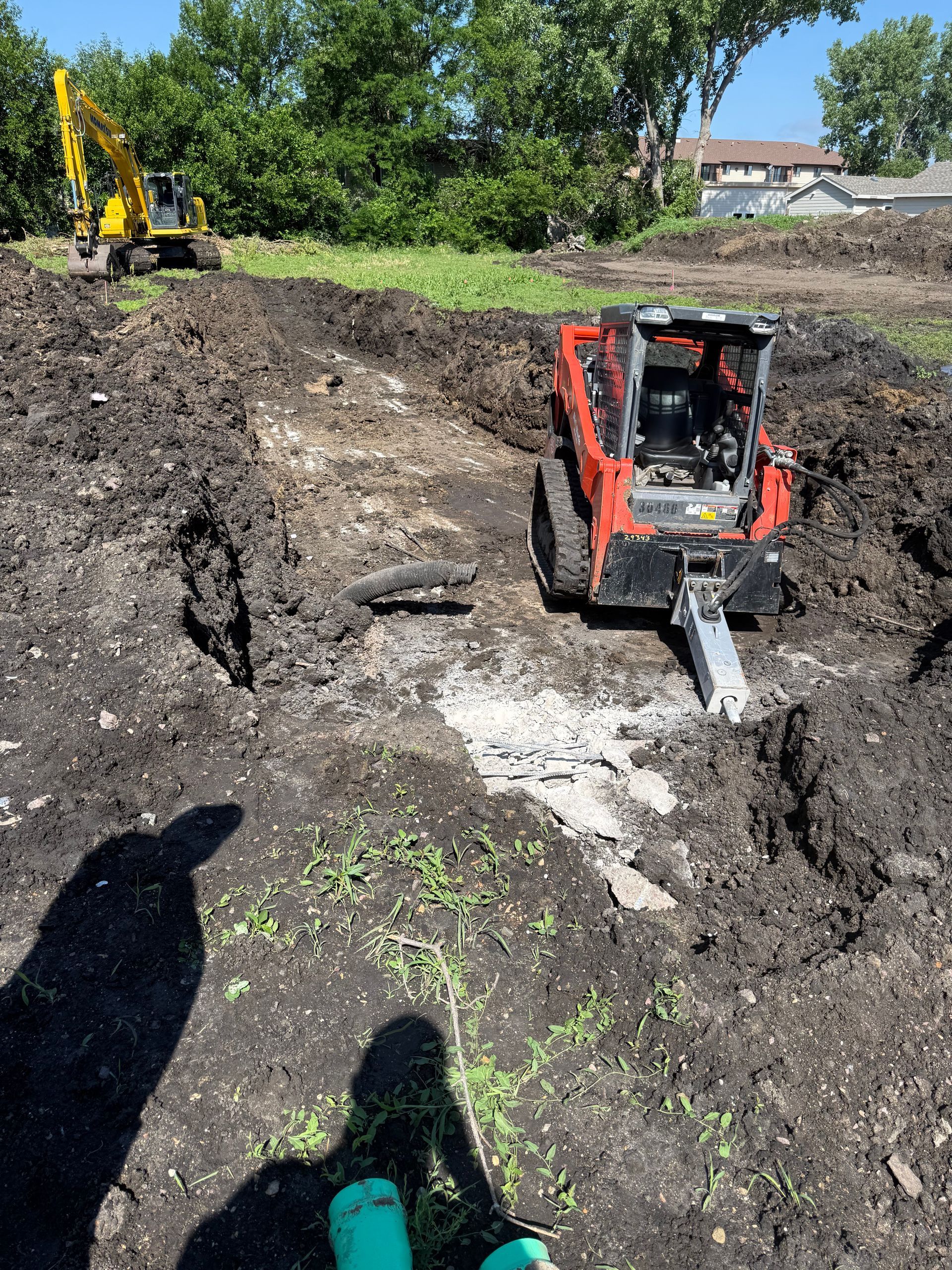 Excavation site: Red skid steer and yellow excavator dig in dirt under a sunny sky.