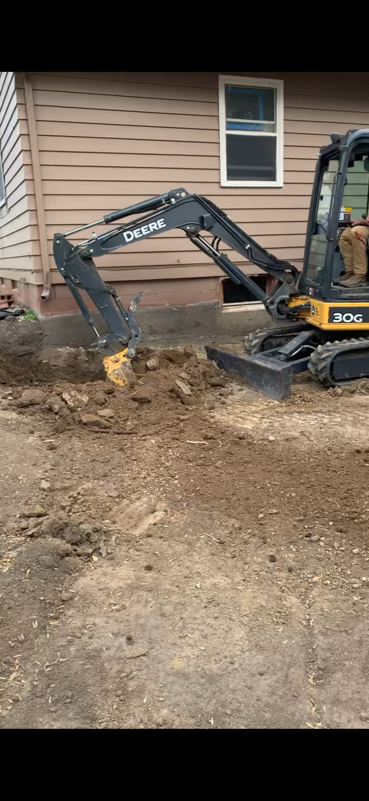 A small John Deere excavator digging dirt next to a tan house.