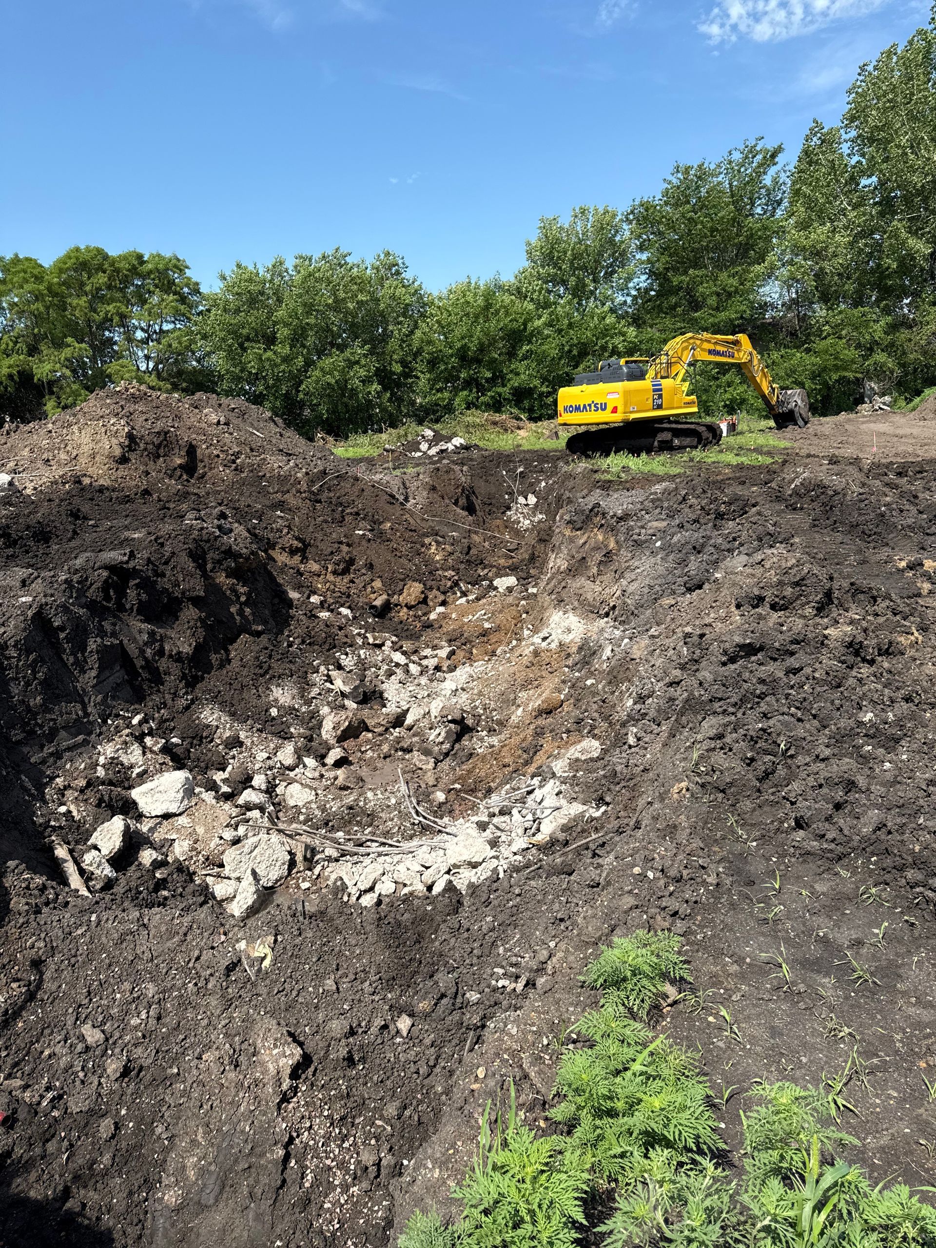 An excavator digging in a dirt pile, revealing rocks and dark soil. Trees and blue sky in background.