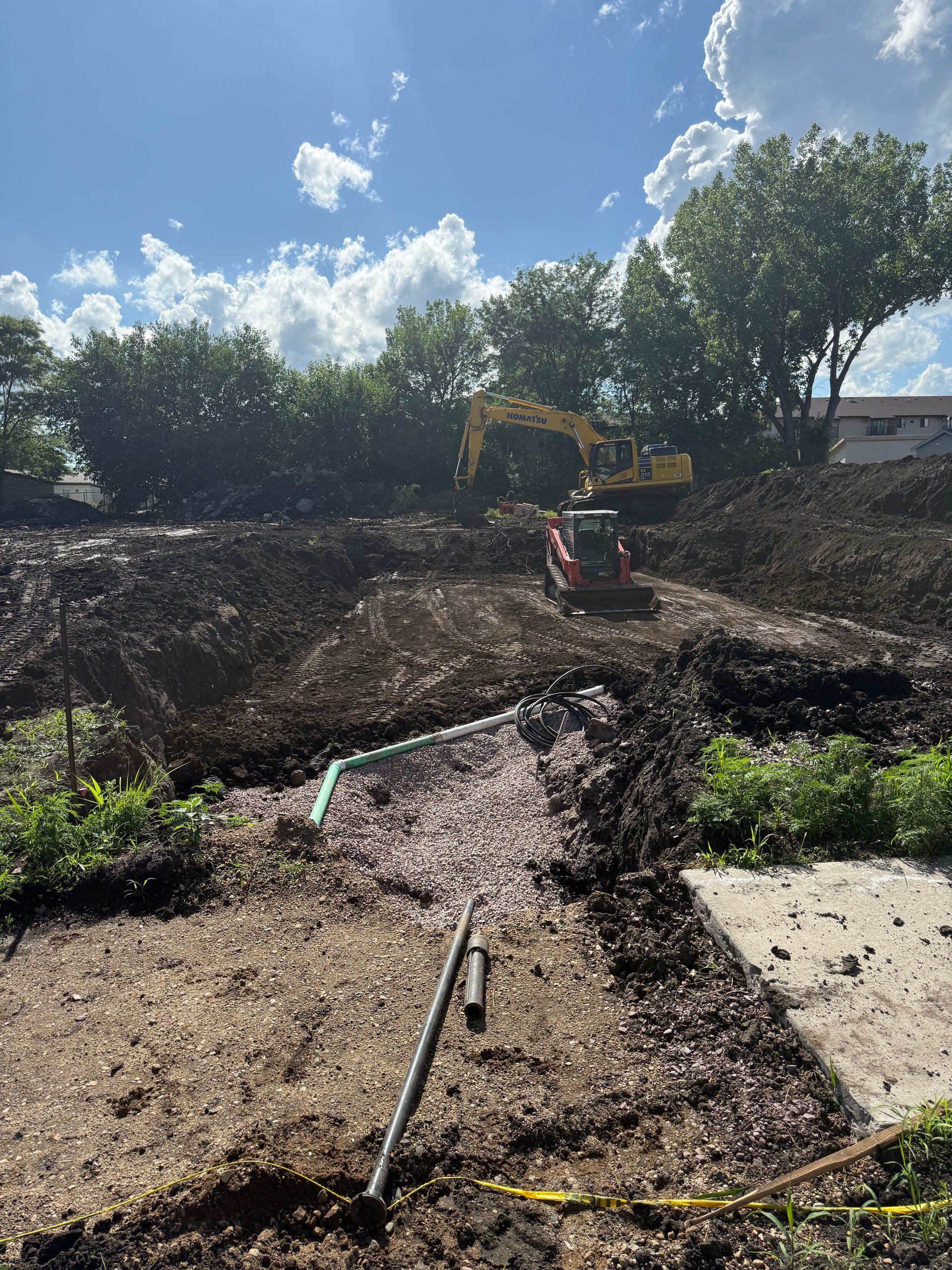 Construction site with excavator and small digger on a dirt hill, green pipe and a clear blue sky.
