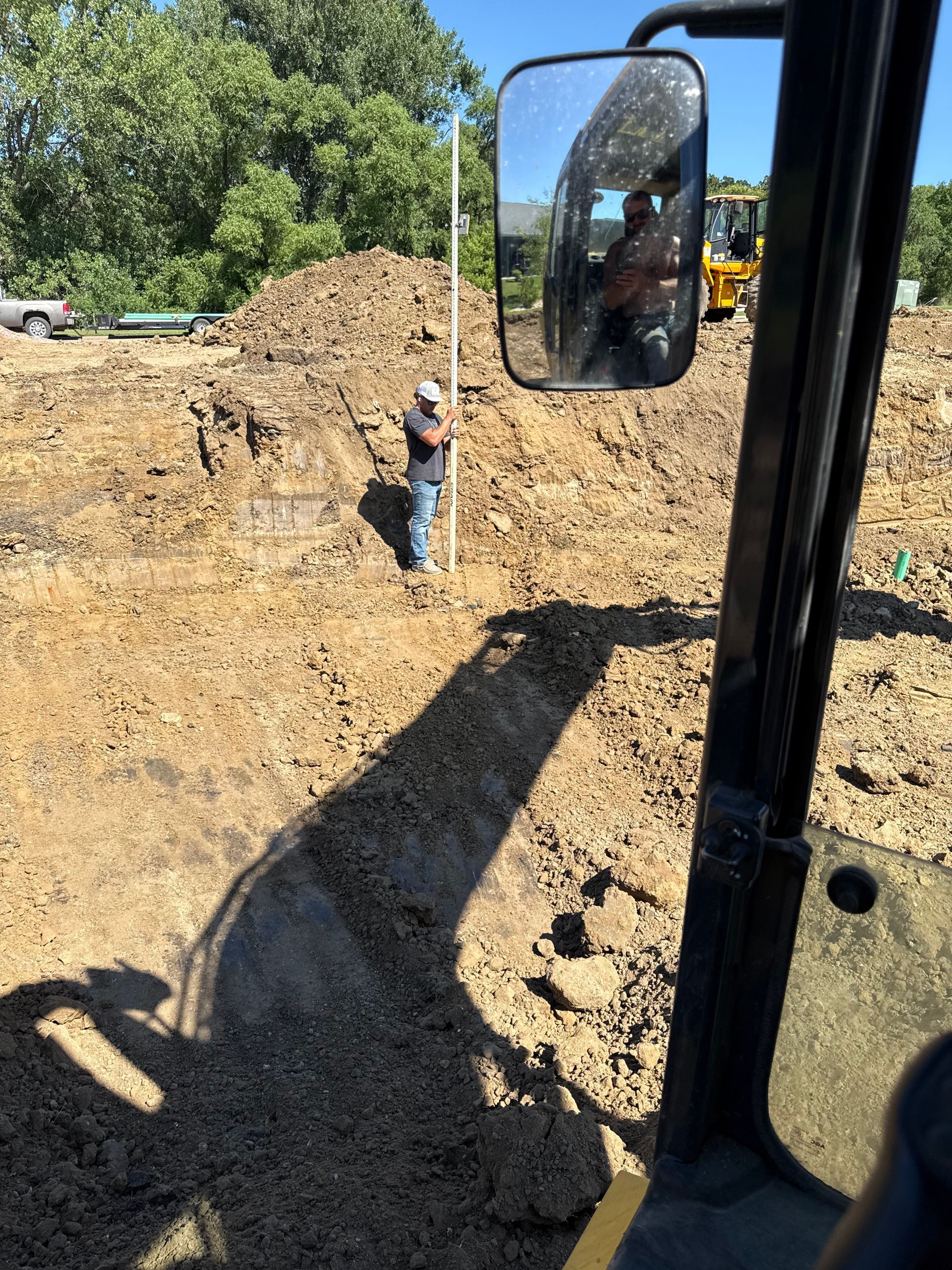 Construction site with person standing near a white pole. Dirt and excavation equipment present, view from inside a machine.