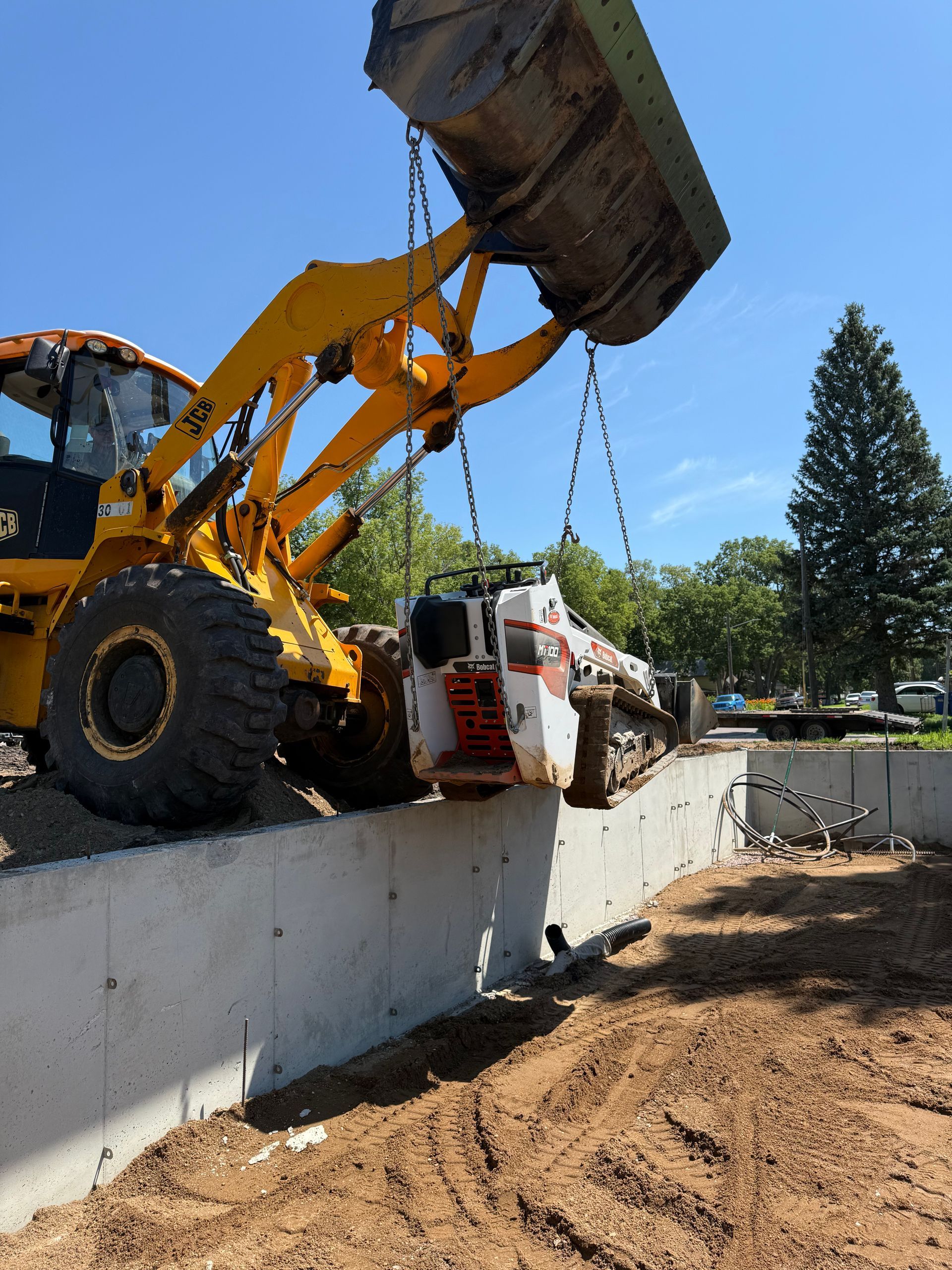 Yellow backhoe lifting a white Bobcat over a concrete wall at a construction site. Sunny, outdoor setting.