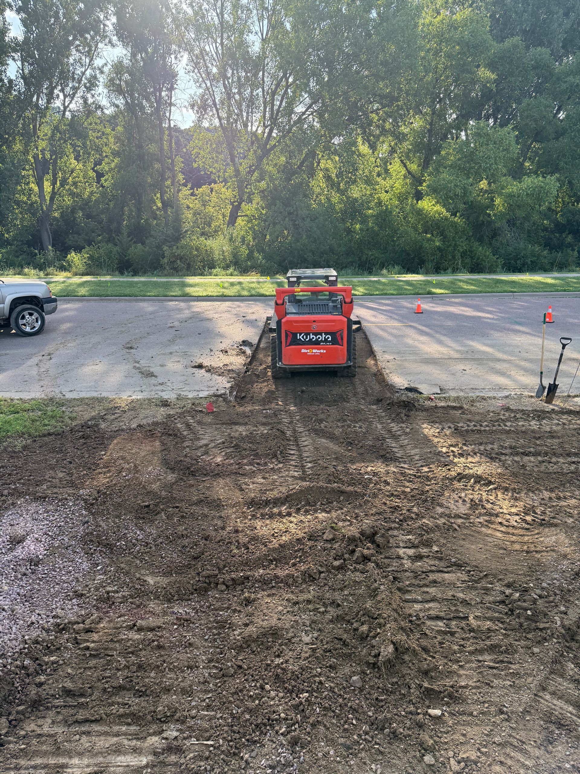 Orange Kubota skid steer on dirt, grading a driveway. Trees and car in background.