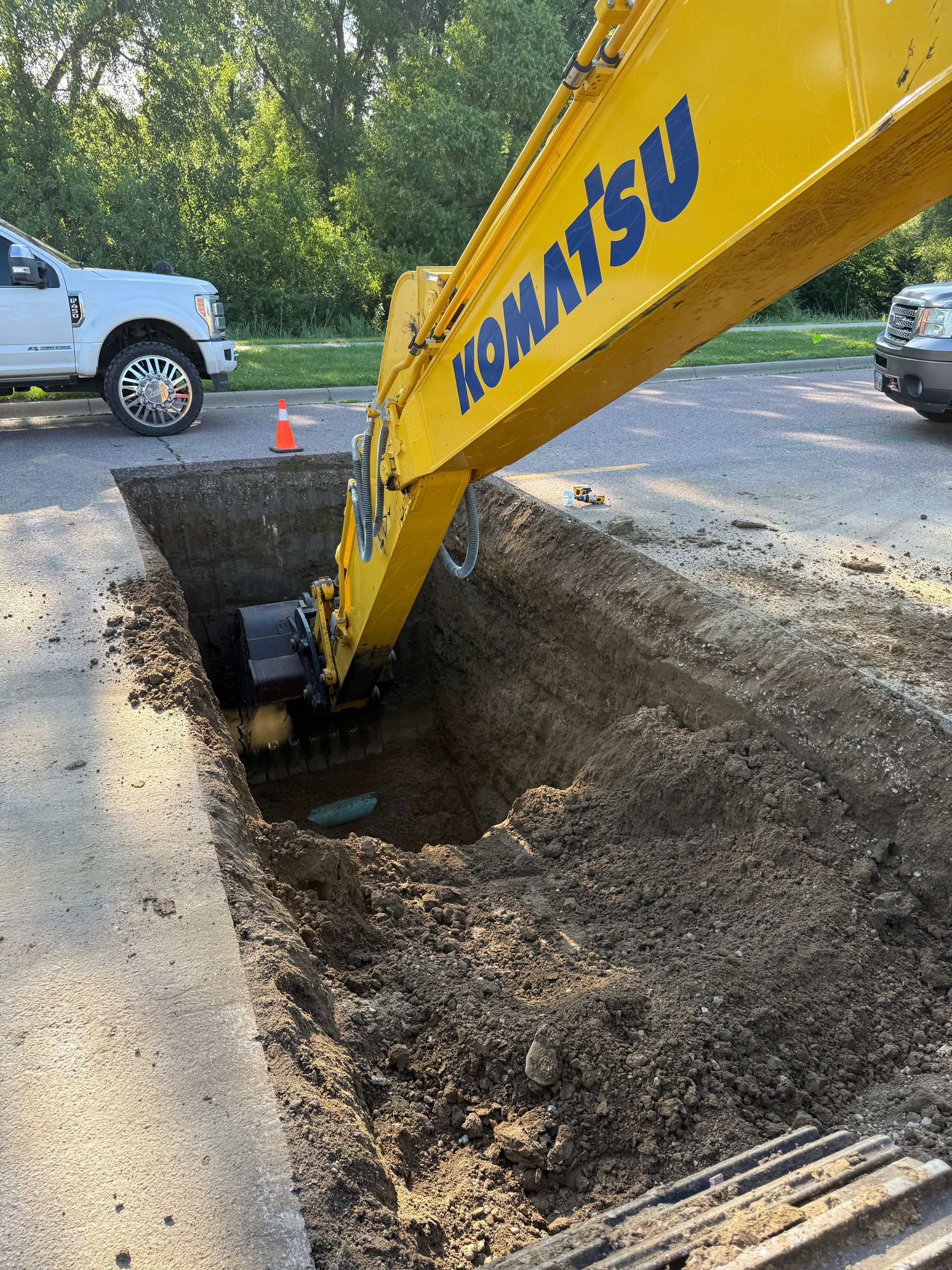 Yellow Komatsu excavator digging a rectangular trench in asphalt road; white truck and parked car nearby.