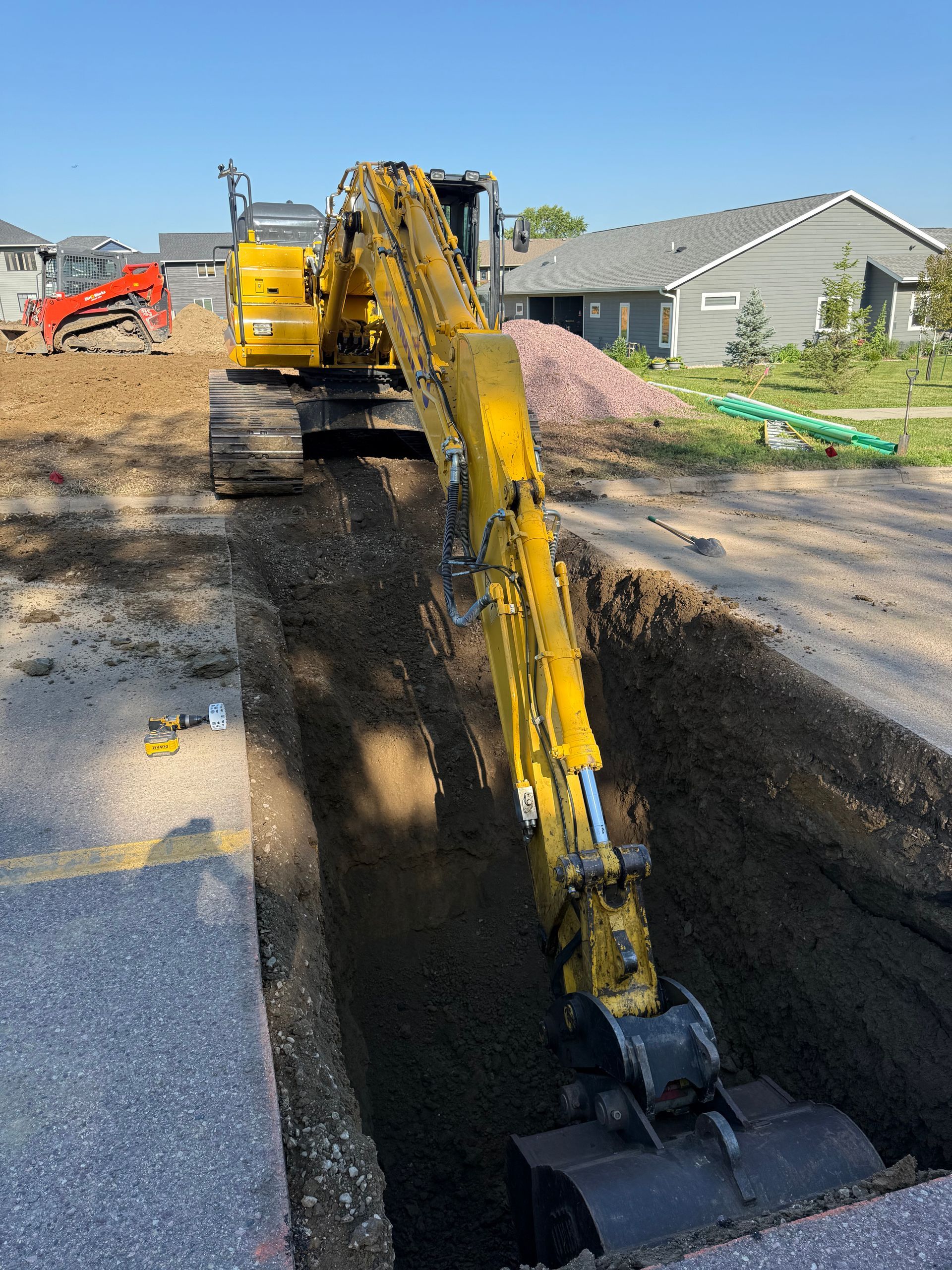 Yellow excavator digging a trench in a street. A small red bulldozer and houses are in the background.