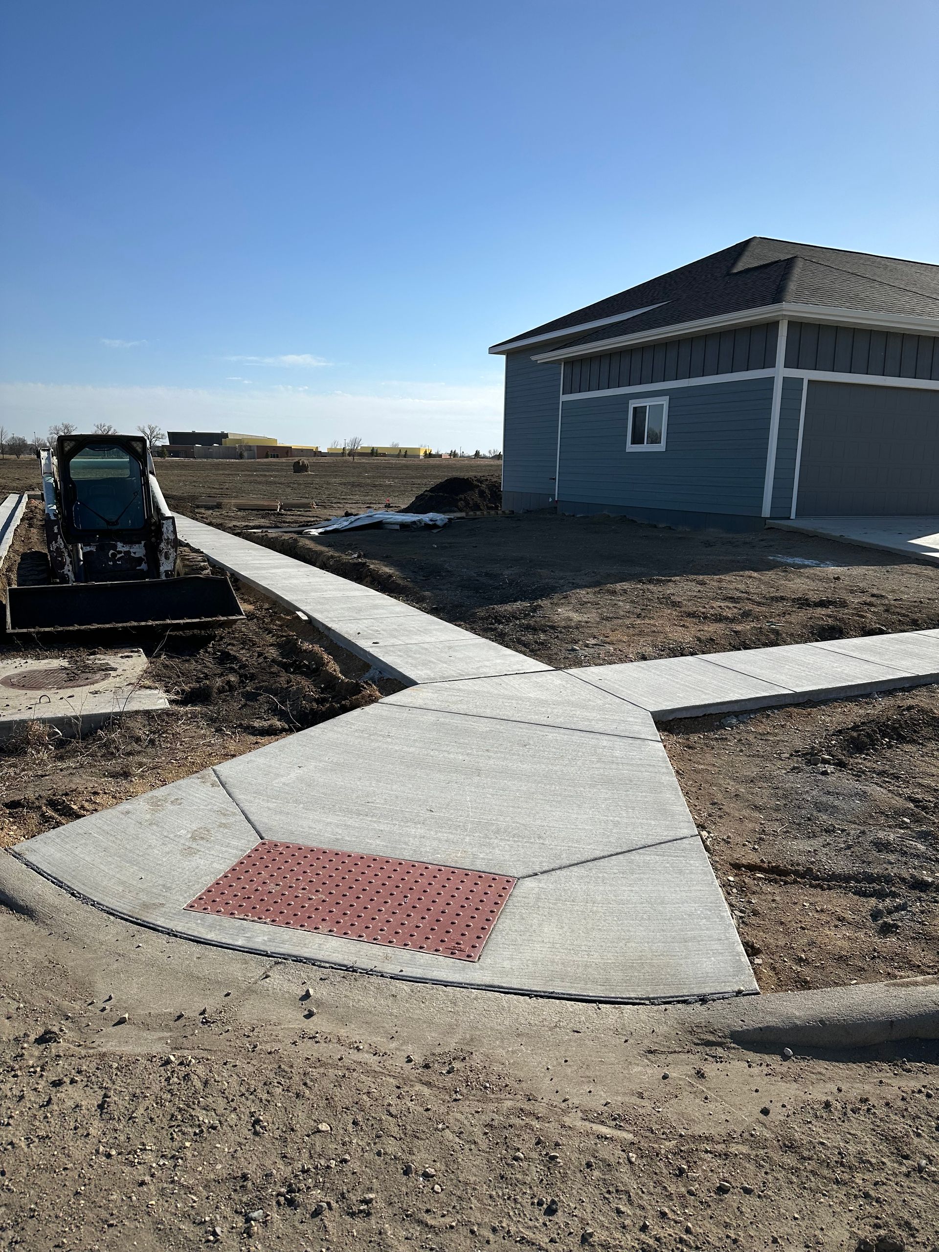 New concrete sidewalks with a red tactile warning tile at a residential intersection; a small skid steer loader sits nearby.