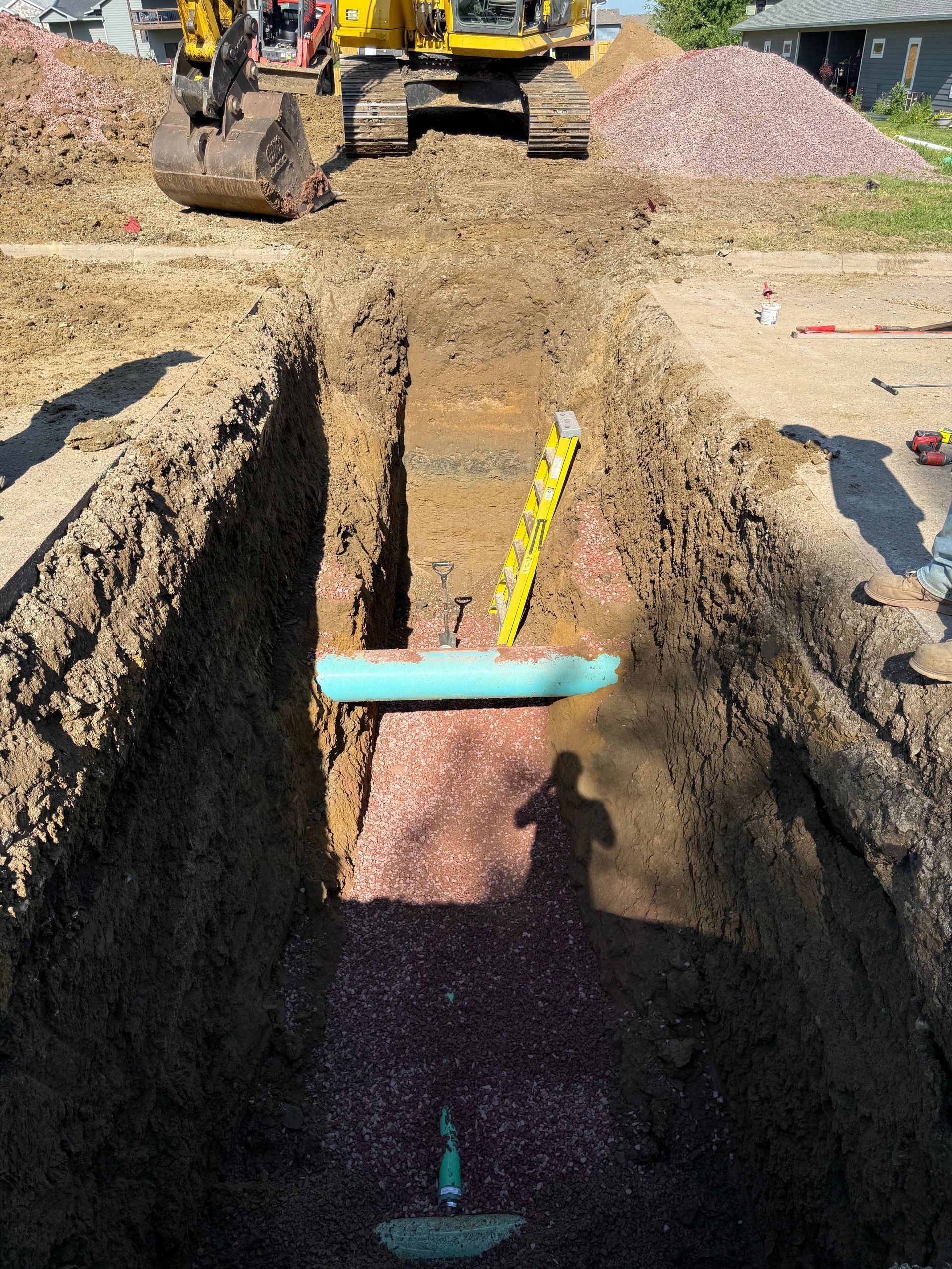 Excavator in trench with exposed pipe. Brown soil, level, pinkish gravel bed.