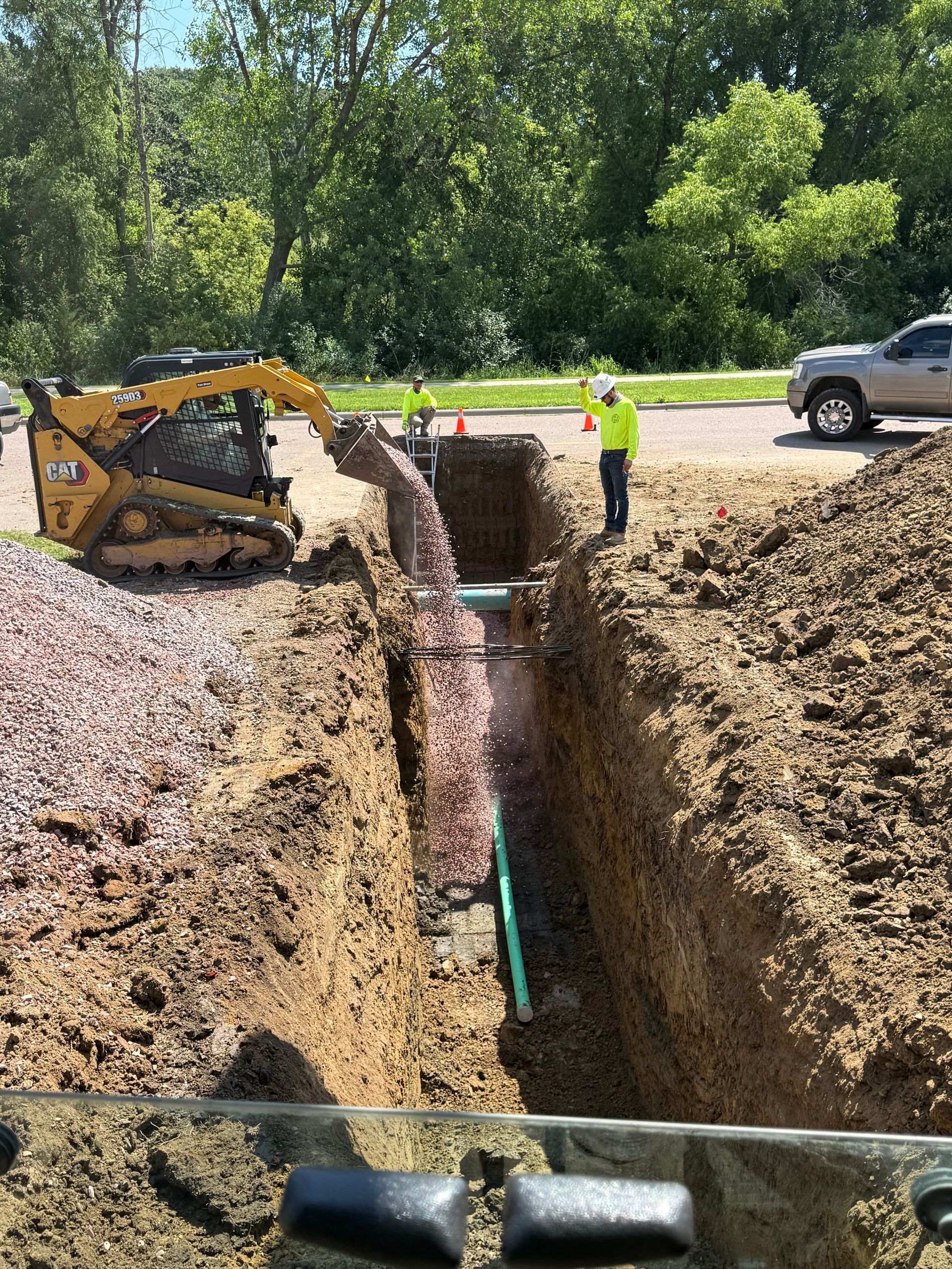 A small construction vehicle adding gravel to a trench holding a green pipe, with a worker observing.
