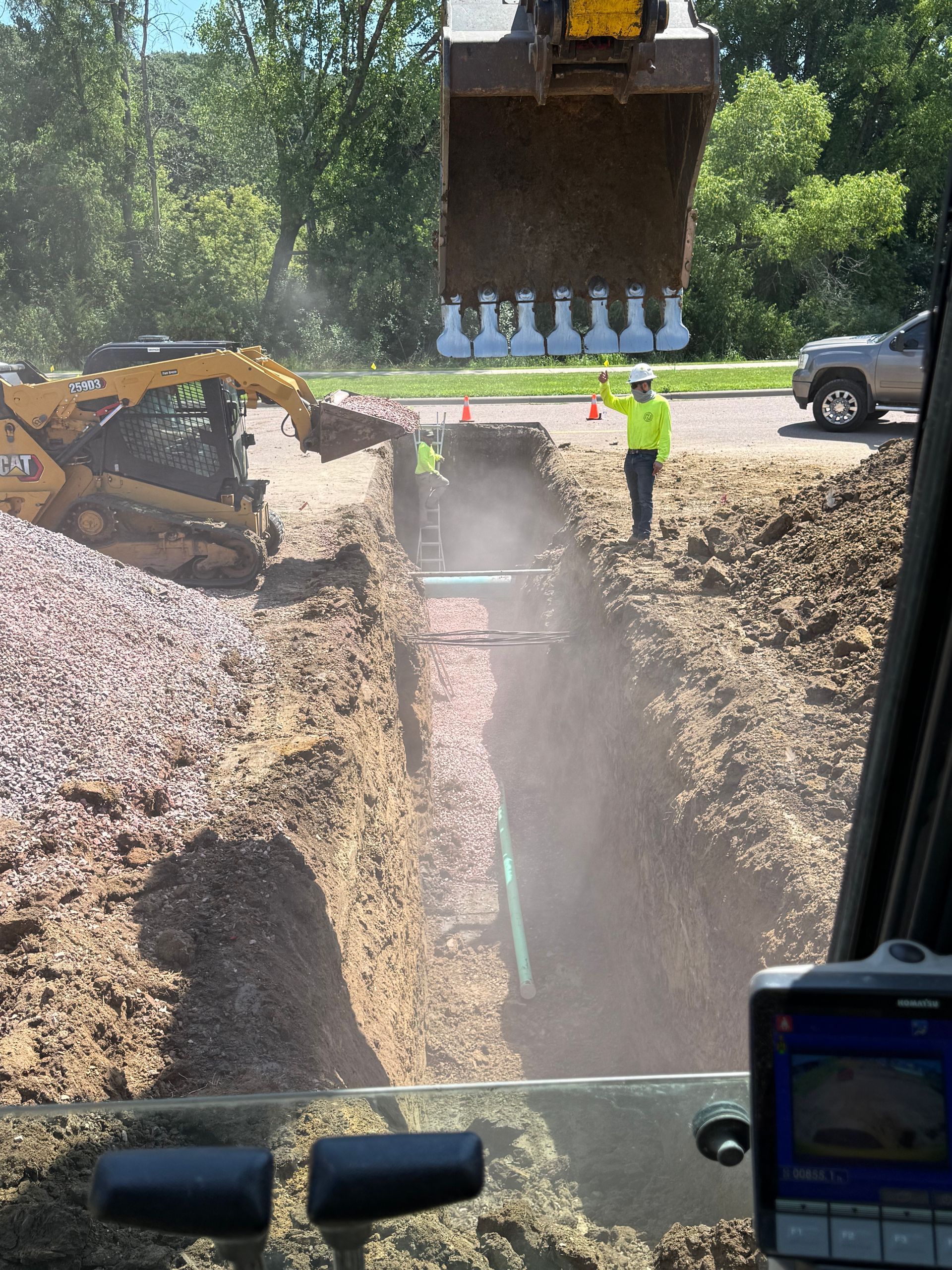 Construction site with an excavator digging a trench, two workers in vests near the trench, a vehicle on the street.