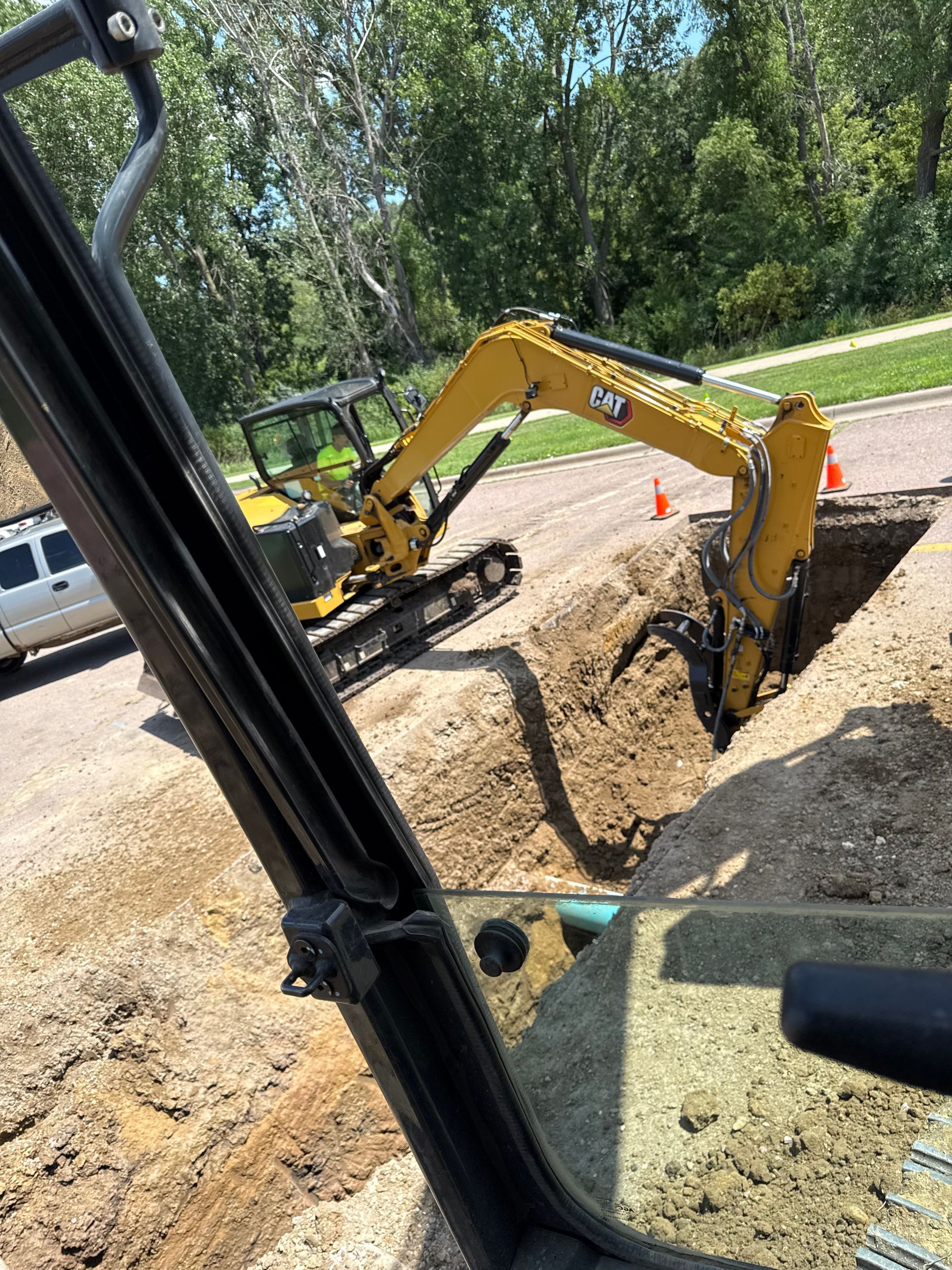 Yellow excavator digging in a trench on a road, trees and a vehicle in the background.