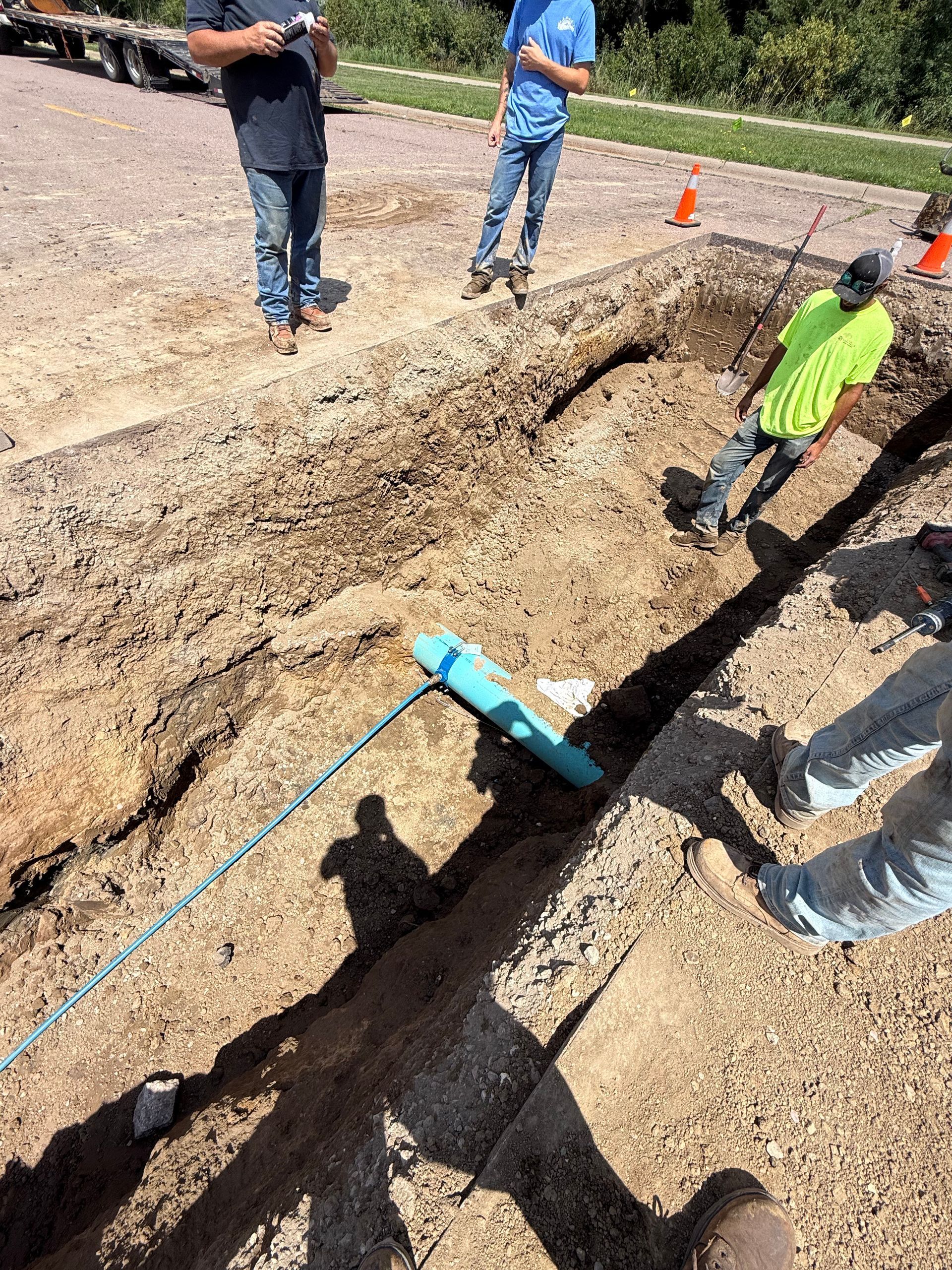Workers installing blue pipe in trench on dirt road. One pulls rope attached to pipe.