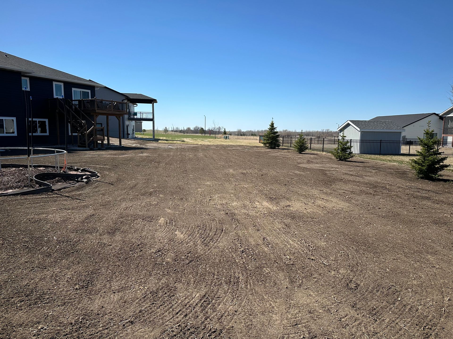 Backyard with dirt, two-story house, small trees, and a clear blue sky.