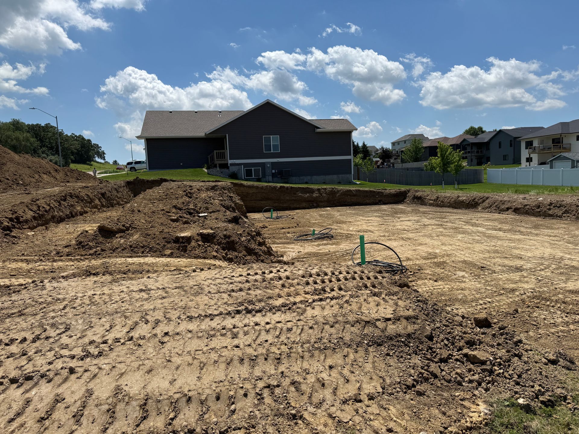 Construction site with a prepared foundation, dirt piles, and a house in the background under a cloudy sky.