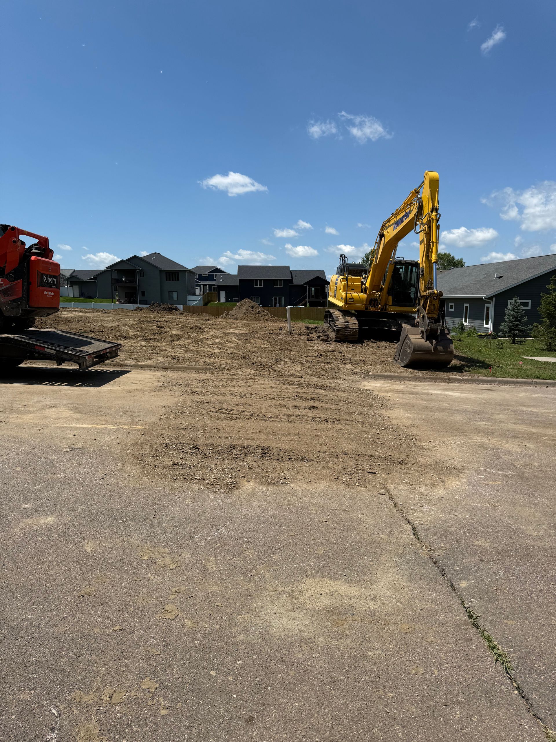 Construction site with excavator and small digger, digging in dirt, houses in background, sunny day.