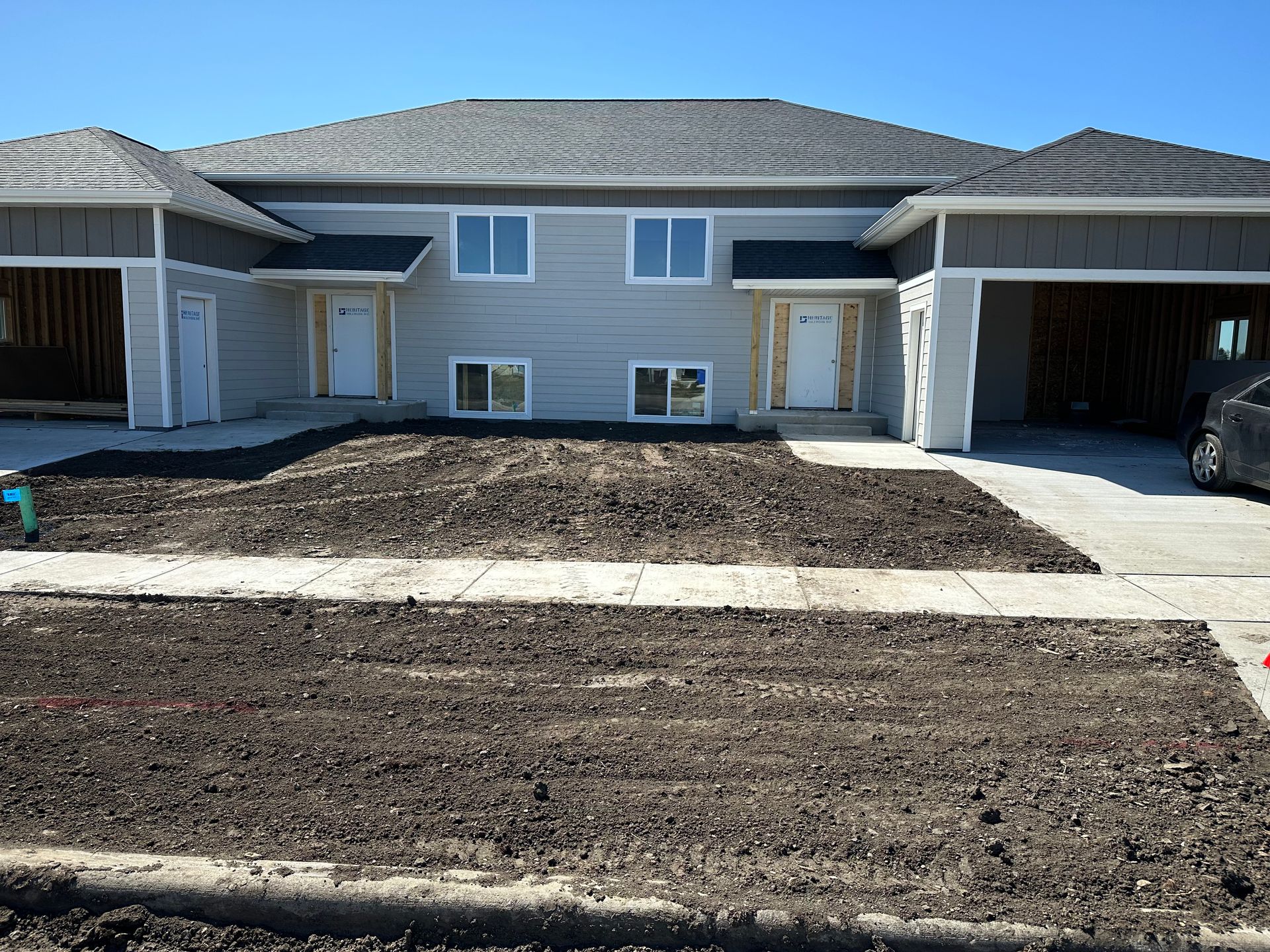 Two-unit, gray house with brown mulch in front yards and open garages; bright blue sky.