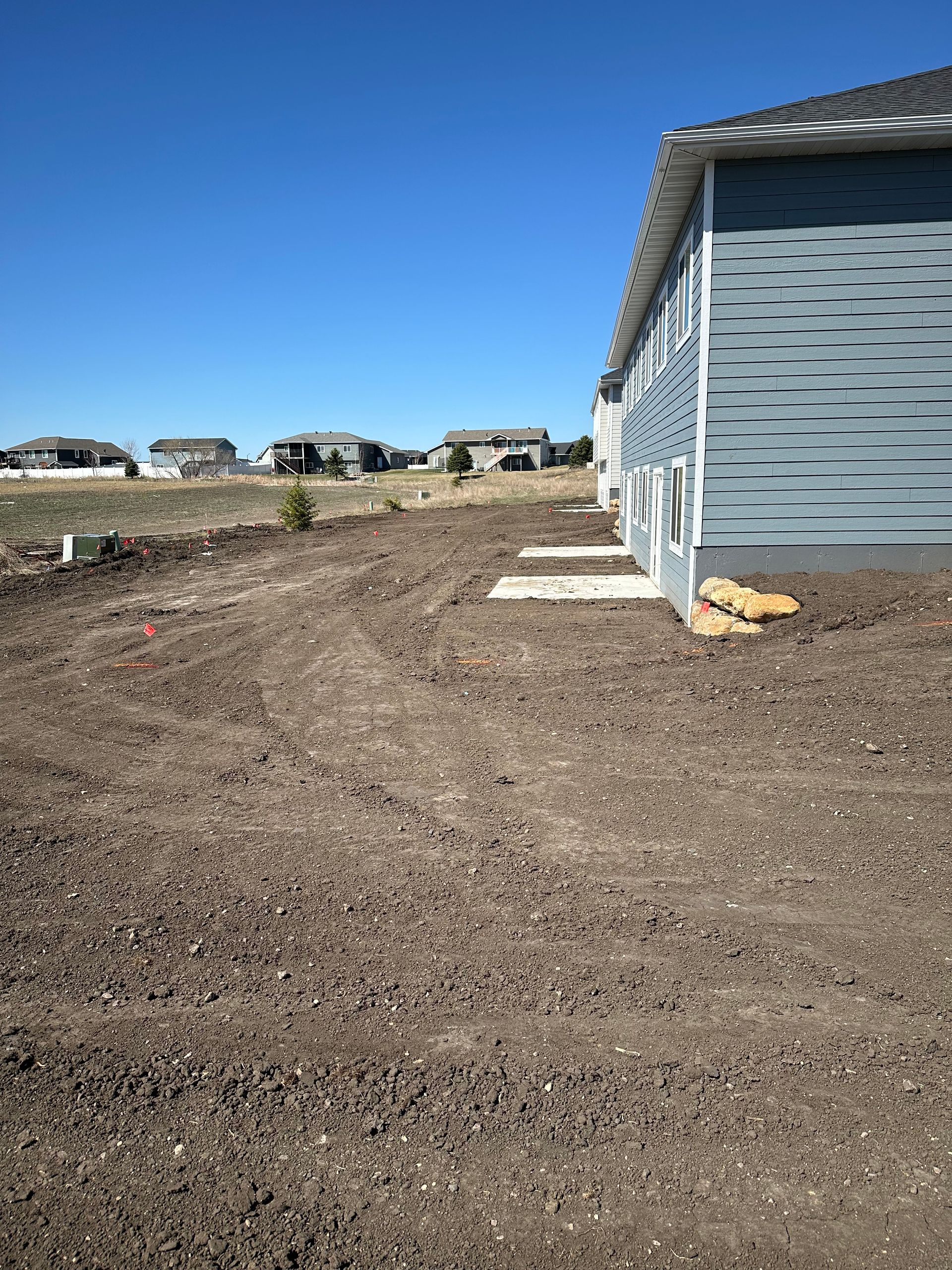 Dirt yard next to a blue building with houses on a hill in the background under a blue sky.
