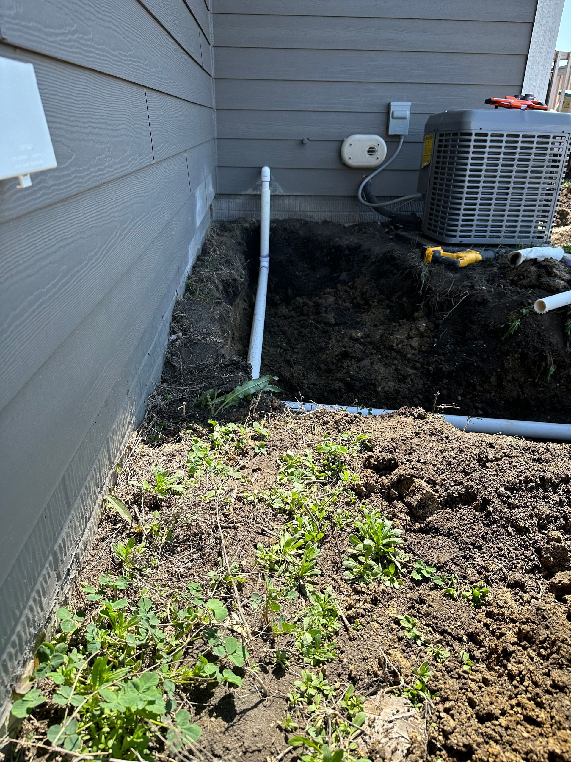 A side view of a house's exterior with dirt and PVC pipes near an air conditioning unit.