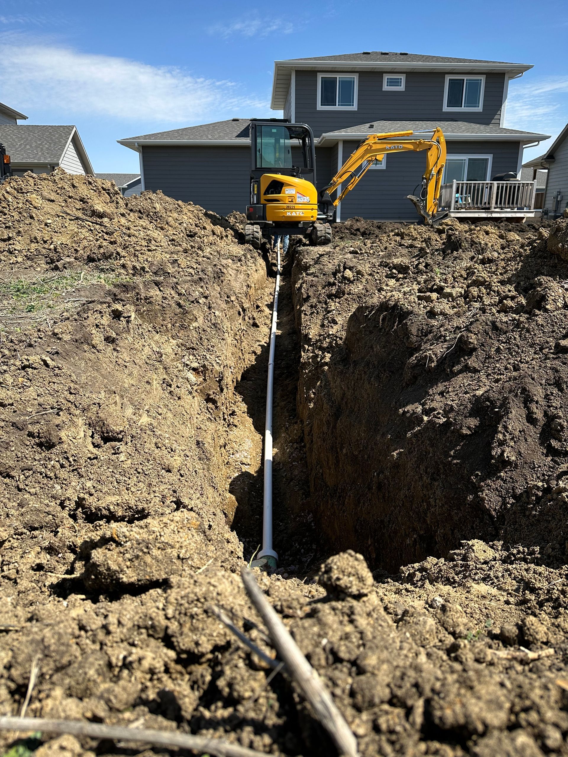 Excavator digging a trench for underground utilities near a house on a sunny day.