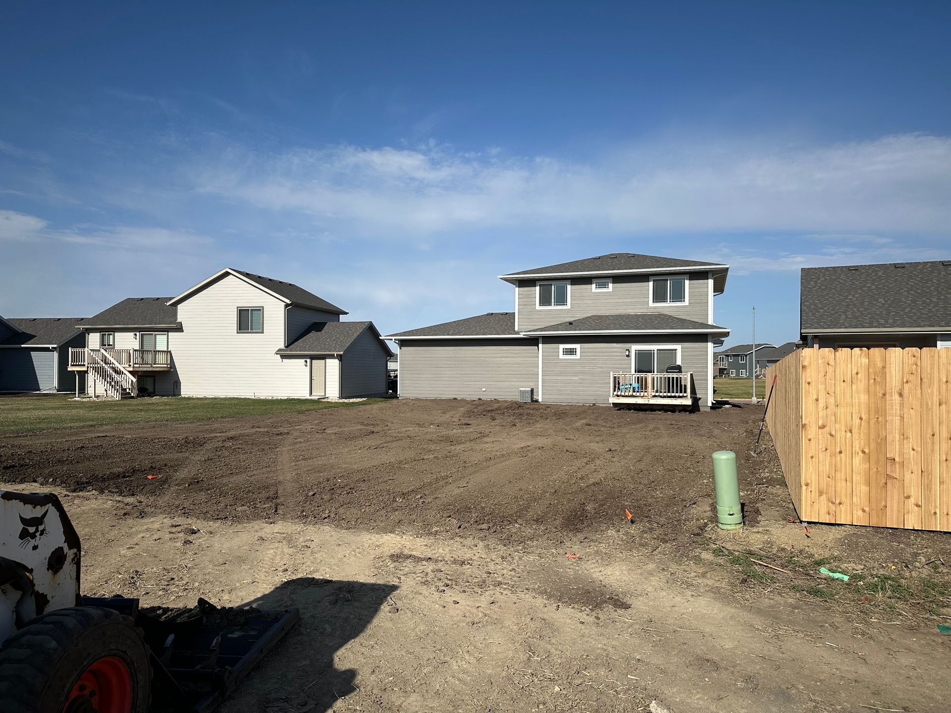 Dirt lot in front of two-story gray house under construction next to a wooden fence and other houses on a sunny day.
