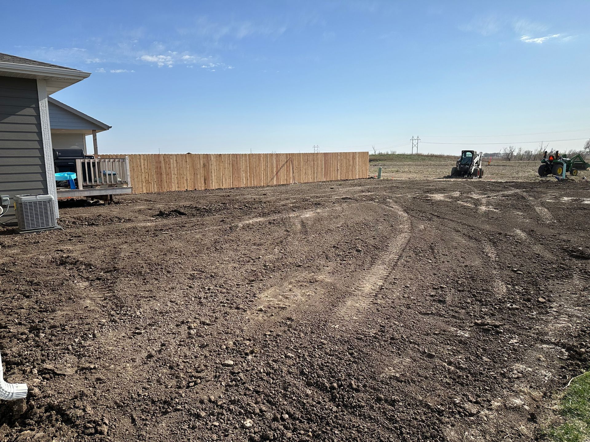 A bare, tilled yard in front of a house, with a wooden fence and tractors in the distance.