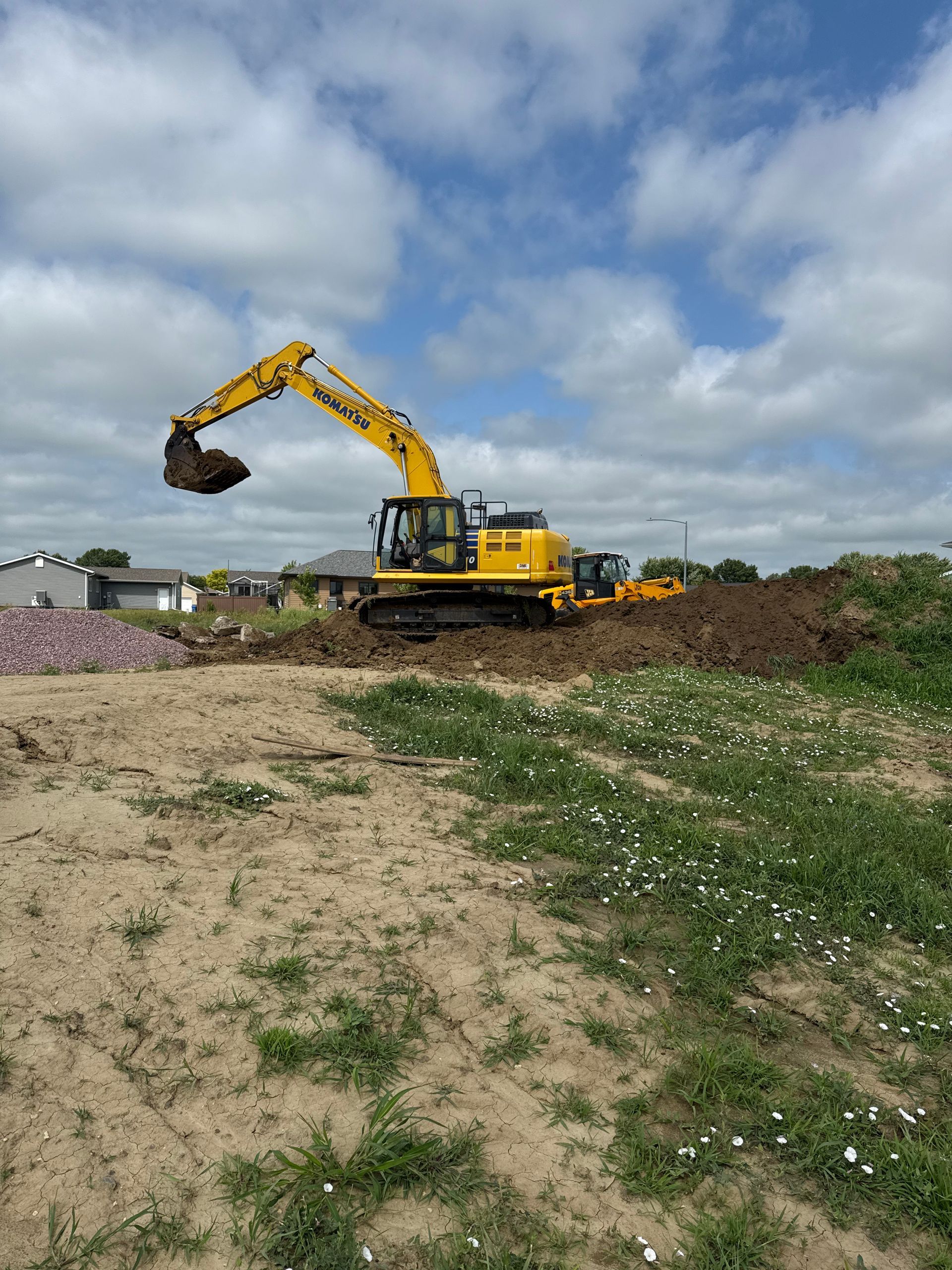 Yellow excavator moving dirt under a cloudy blue sky. A bulldozer is also present.