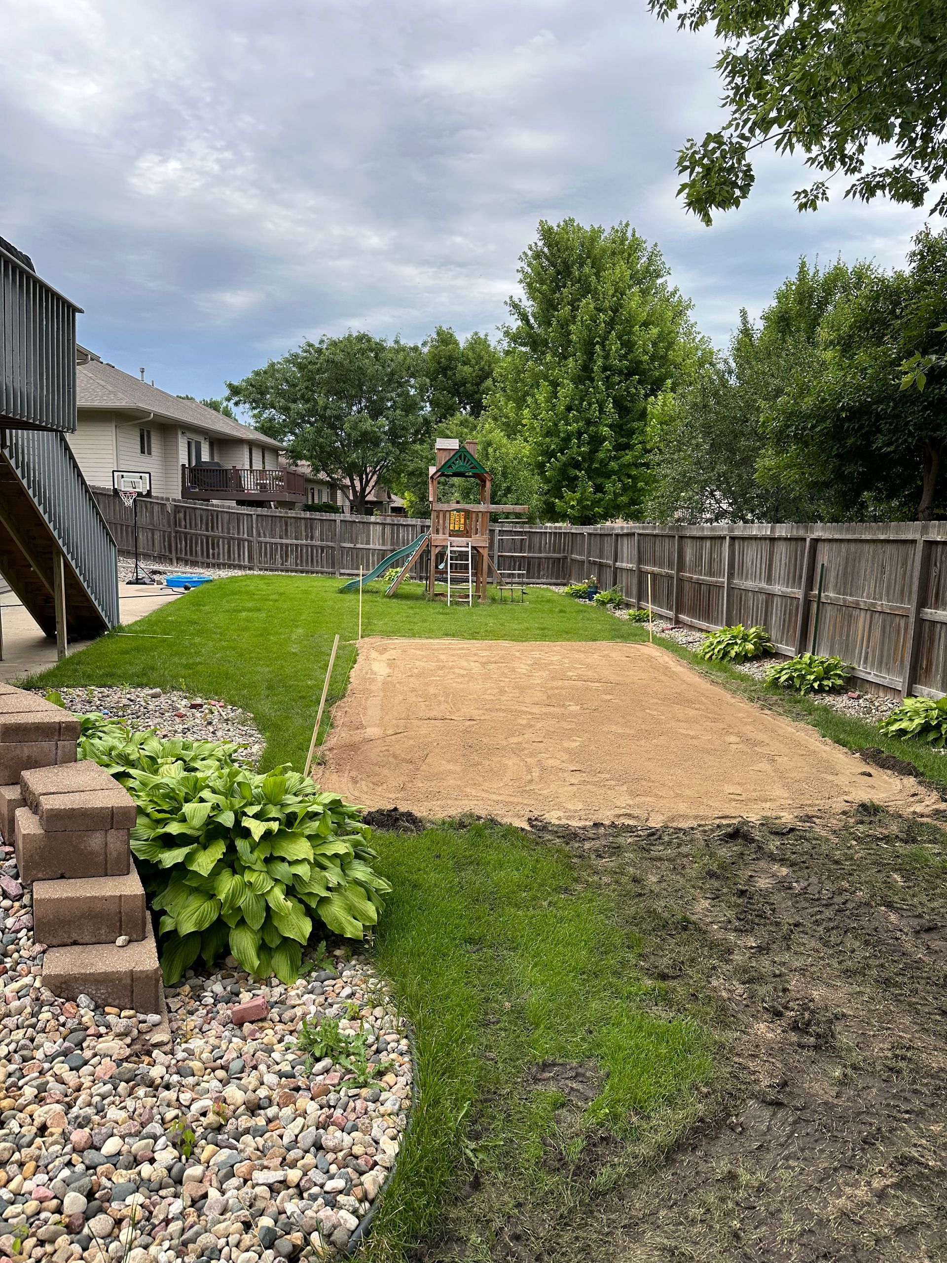 Backyard with a dirt patch, grass, a wooden fence, and a play structure. Overcast sky.