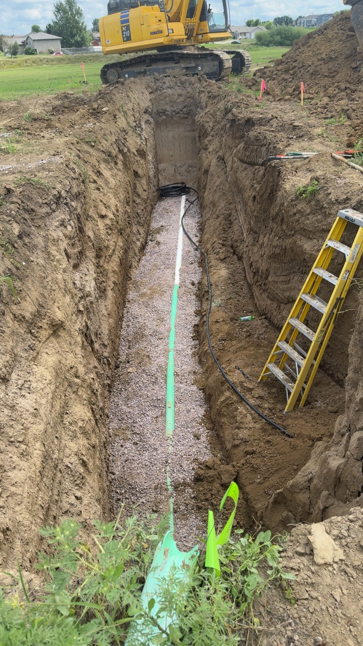 Trench with gravel and pipe, construction site. Excavator in background, ladder in the trench.