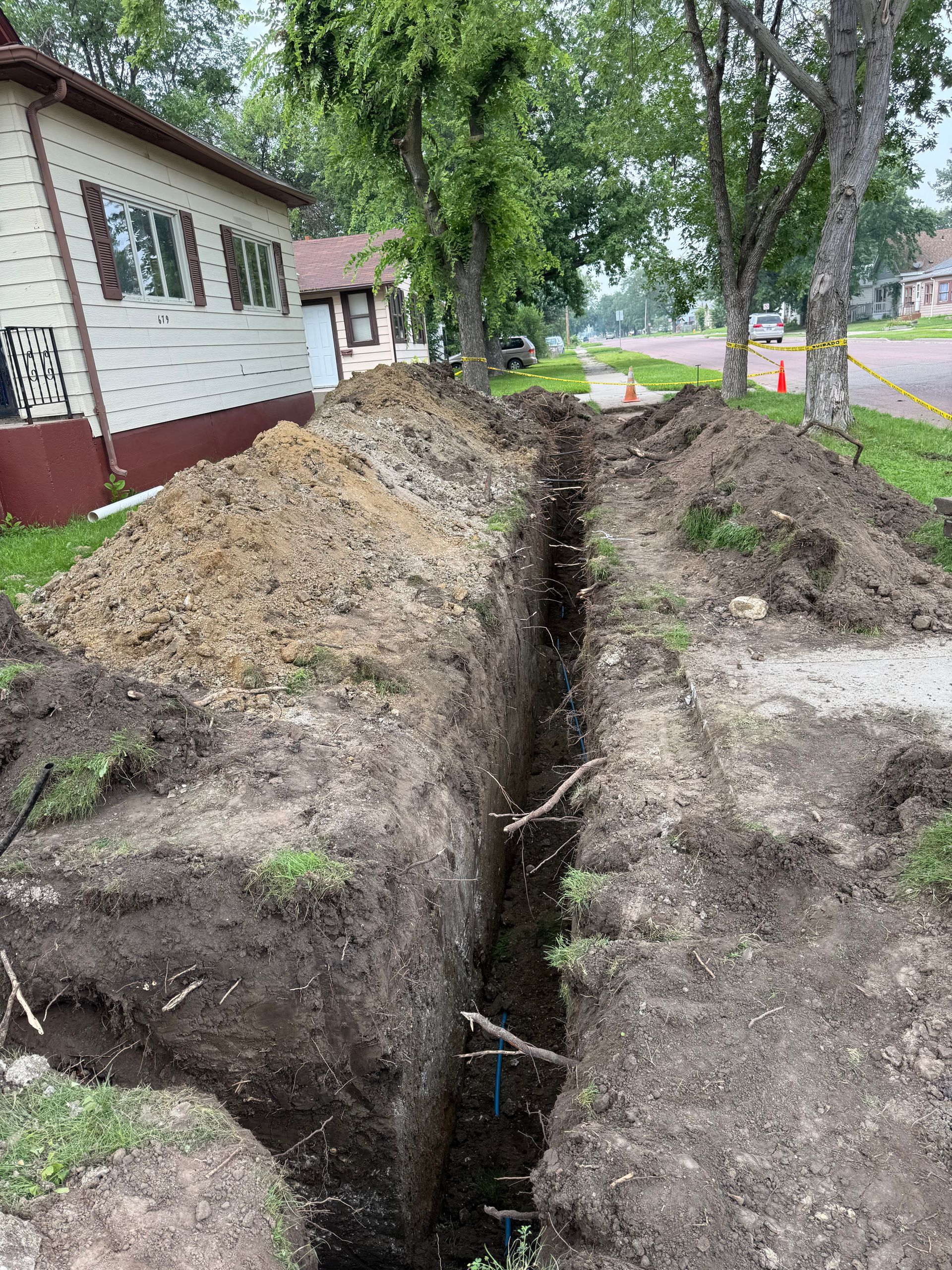 Trench dug alongside a sidewalk, running between a house and street. Dirt pile to the side.