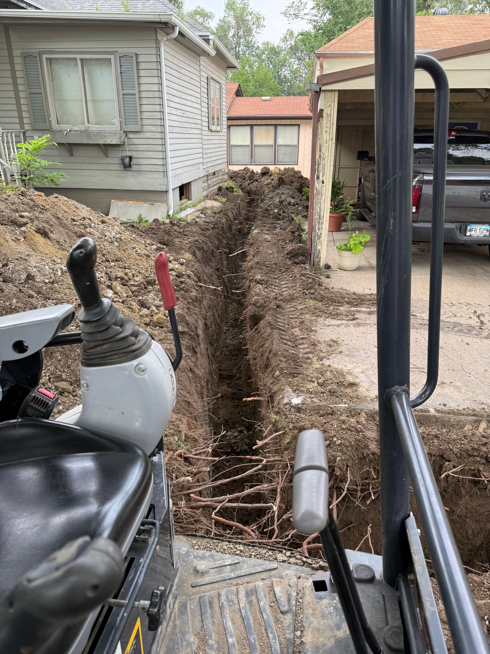 Mini excavator digging a trench between two houses. Earth is piled on the left. The sky is overcast.