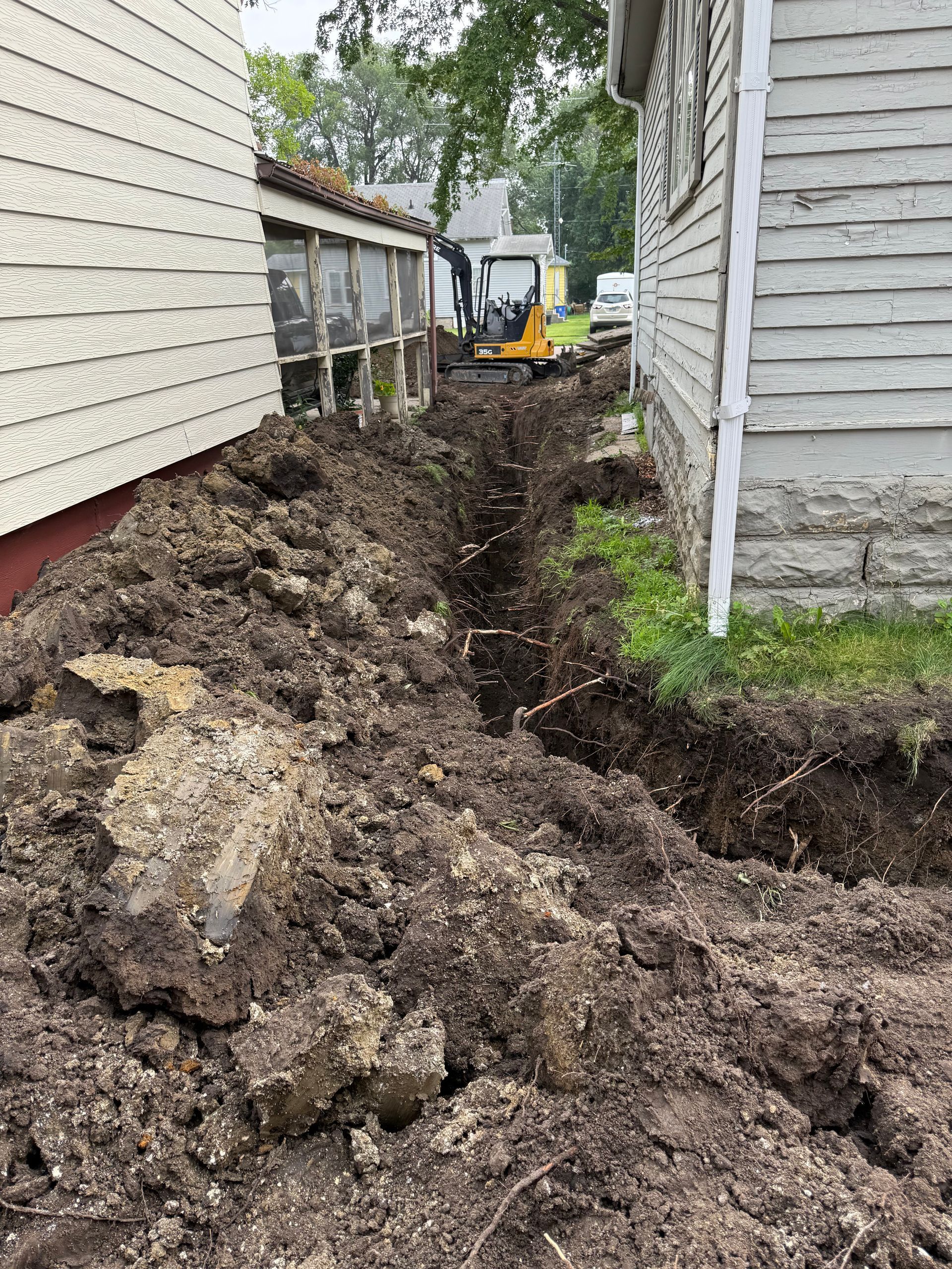 Trench dug between two houses. Dirt pile alongside, mini excavator in the distance, person working.