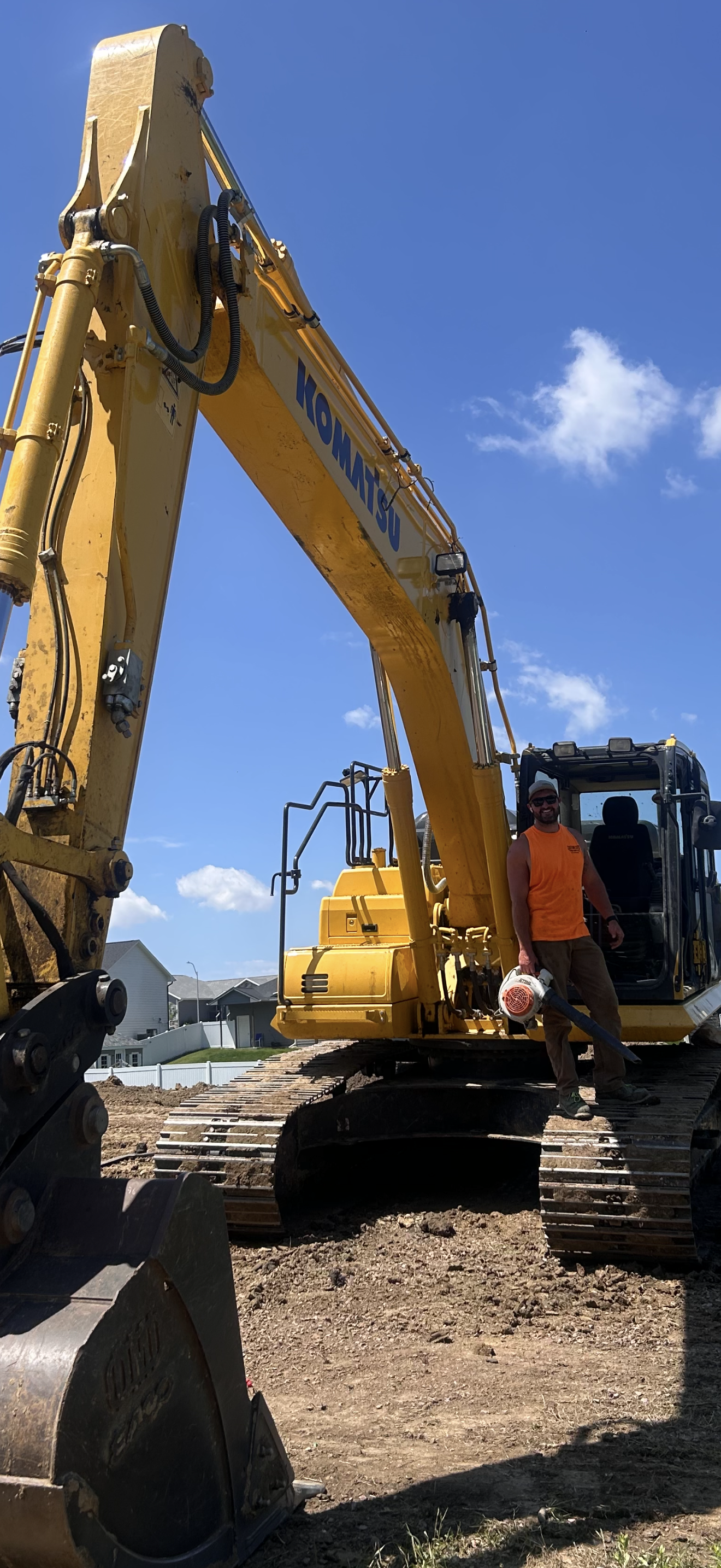 A yellow Komatsu excavator on a construction site with a worker standing near the cab, under a bright blue sky.