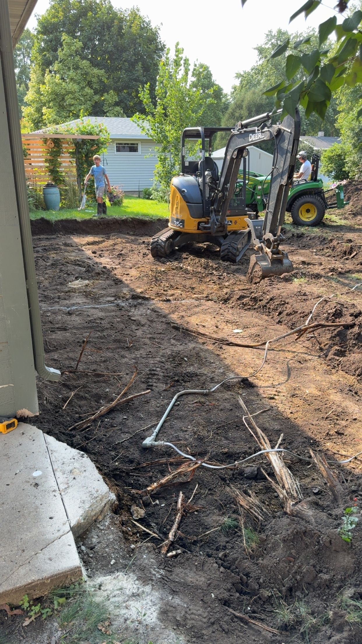 Construction site: Excavator and worker digging in dirt, trees and house in background.