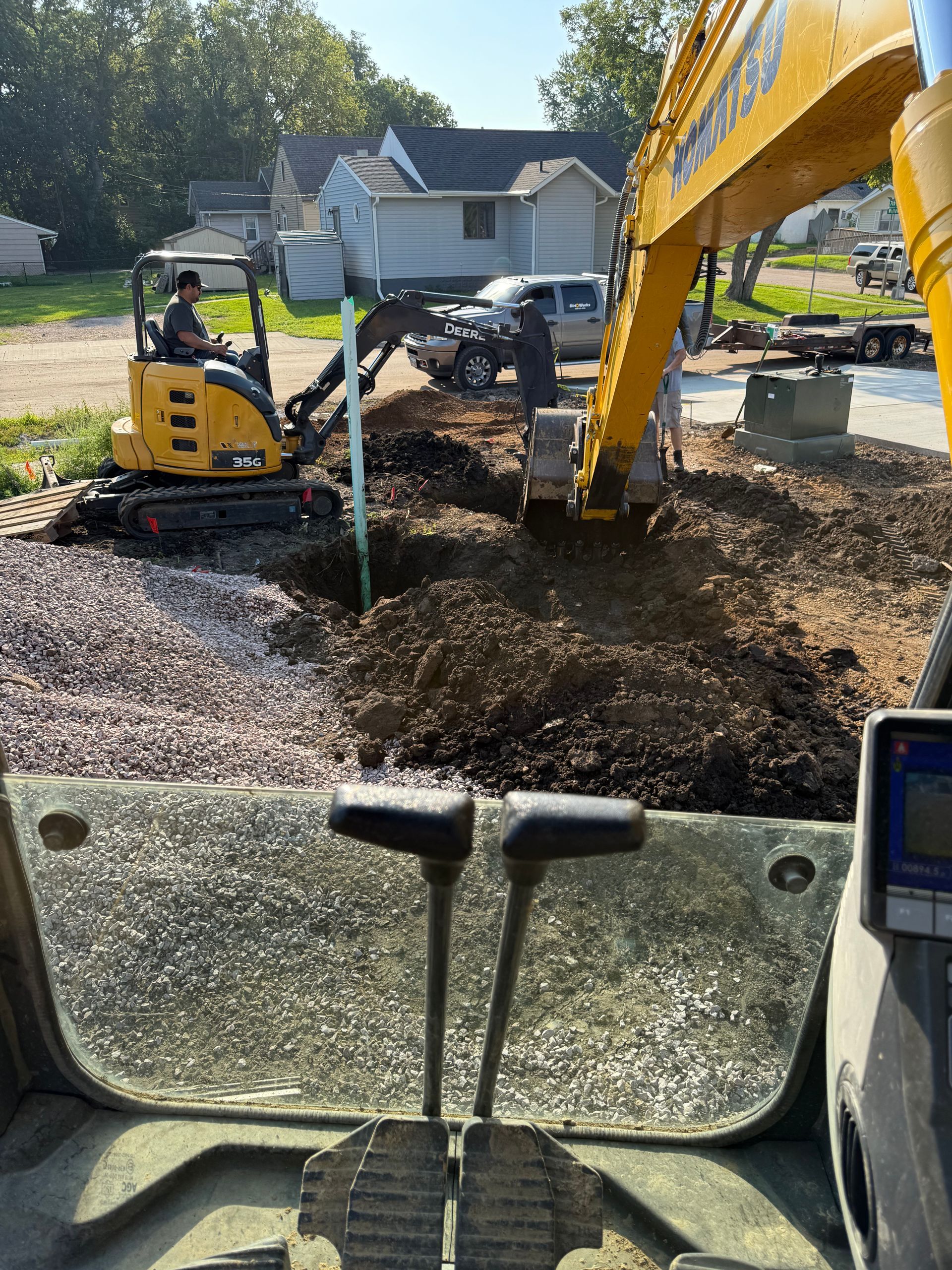 An excavator digs a hole in a residential yard. The operator sits inside the yellow machine.