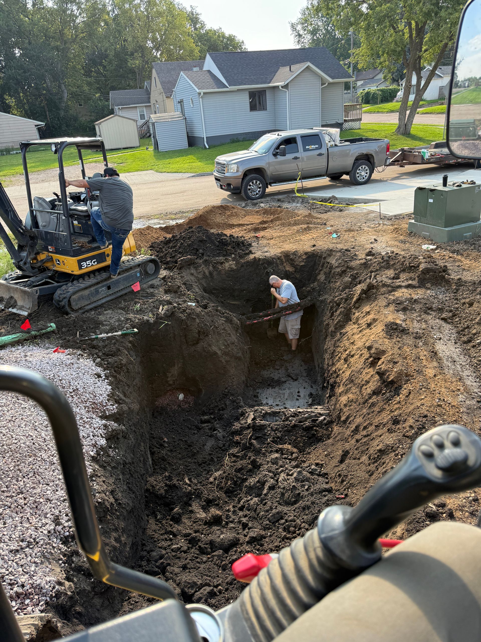 Construction site: Mini excavator digs a hole as a worker in the hole and another operates the machine. A pickup truck is parked in the background.
