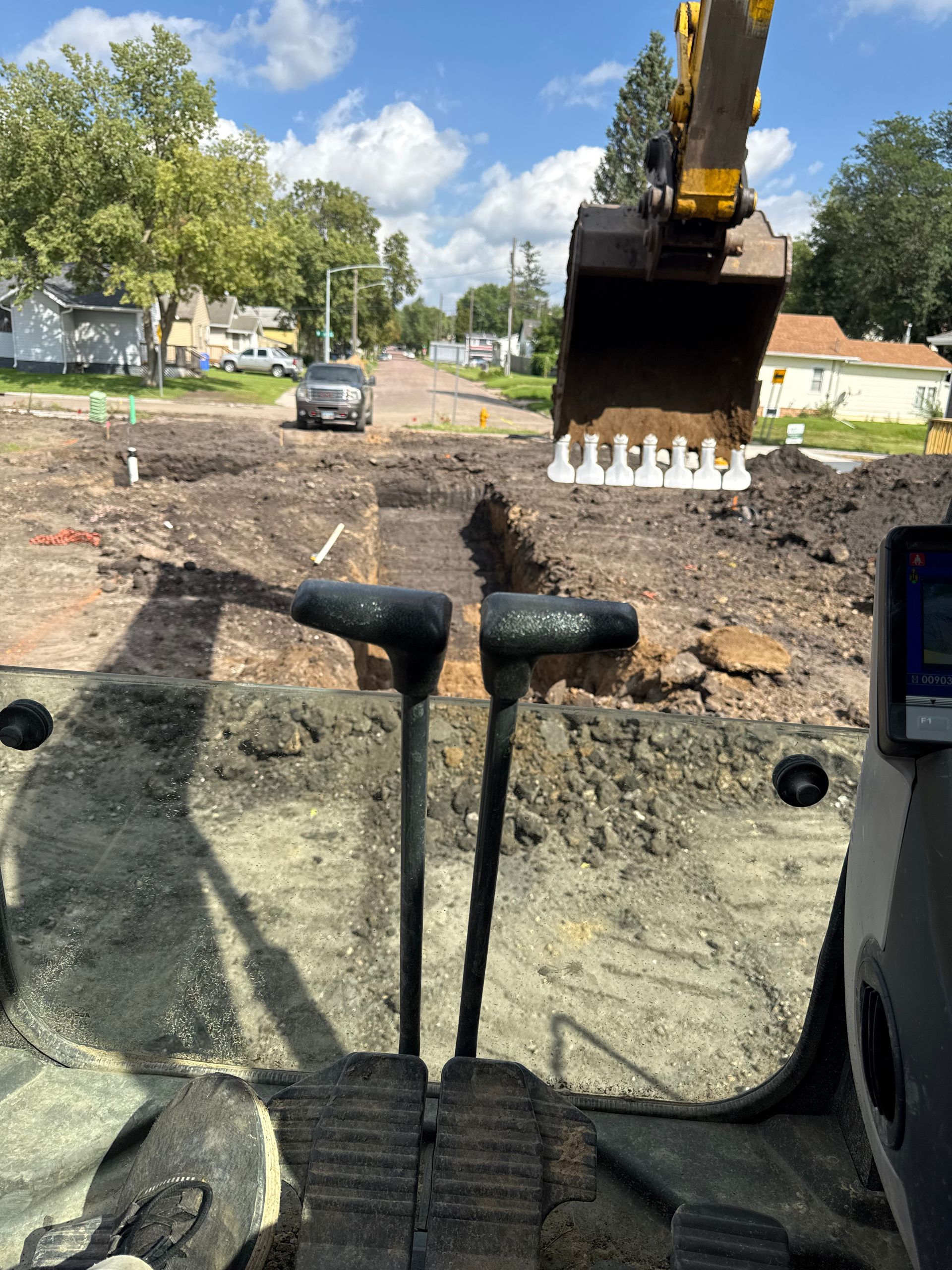 View from excavator cab, digging trench in a road. Sunny day. Road, houses, and car in background.