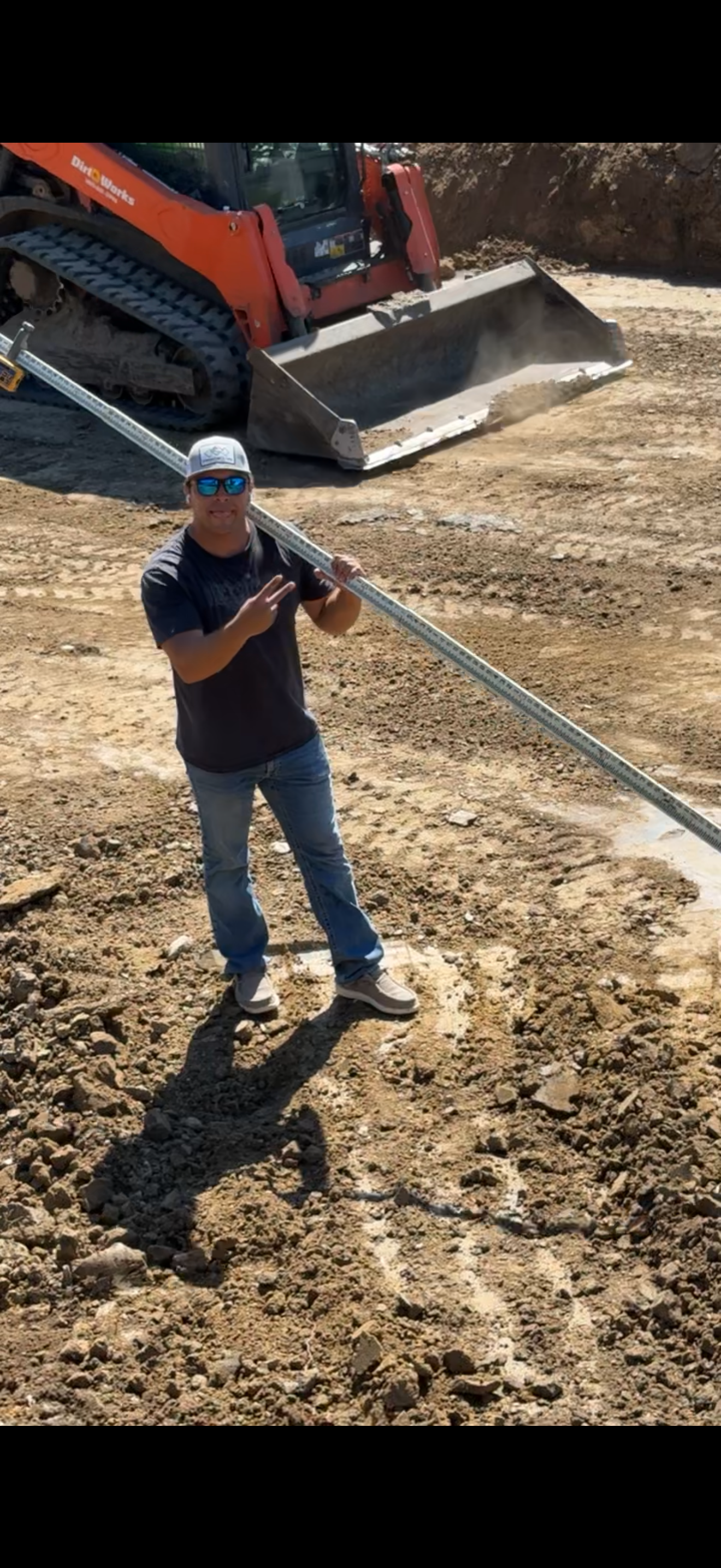 Man pointing at metal pole, standing near construction site and orange skid steer.