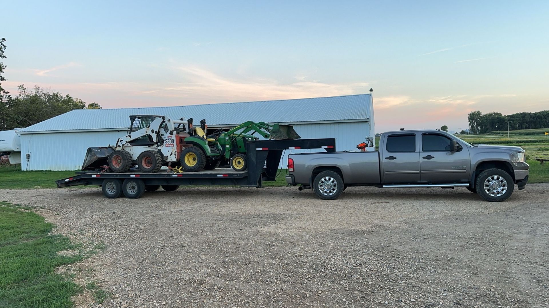 Truck towing a trailer carrying a skid steer loader and a tractor in front of a white building.