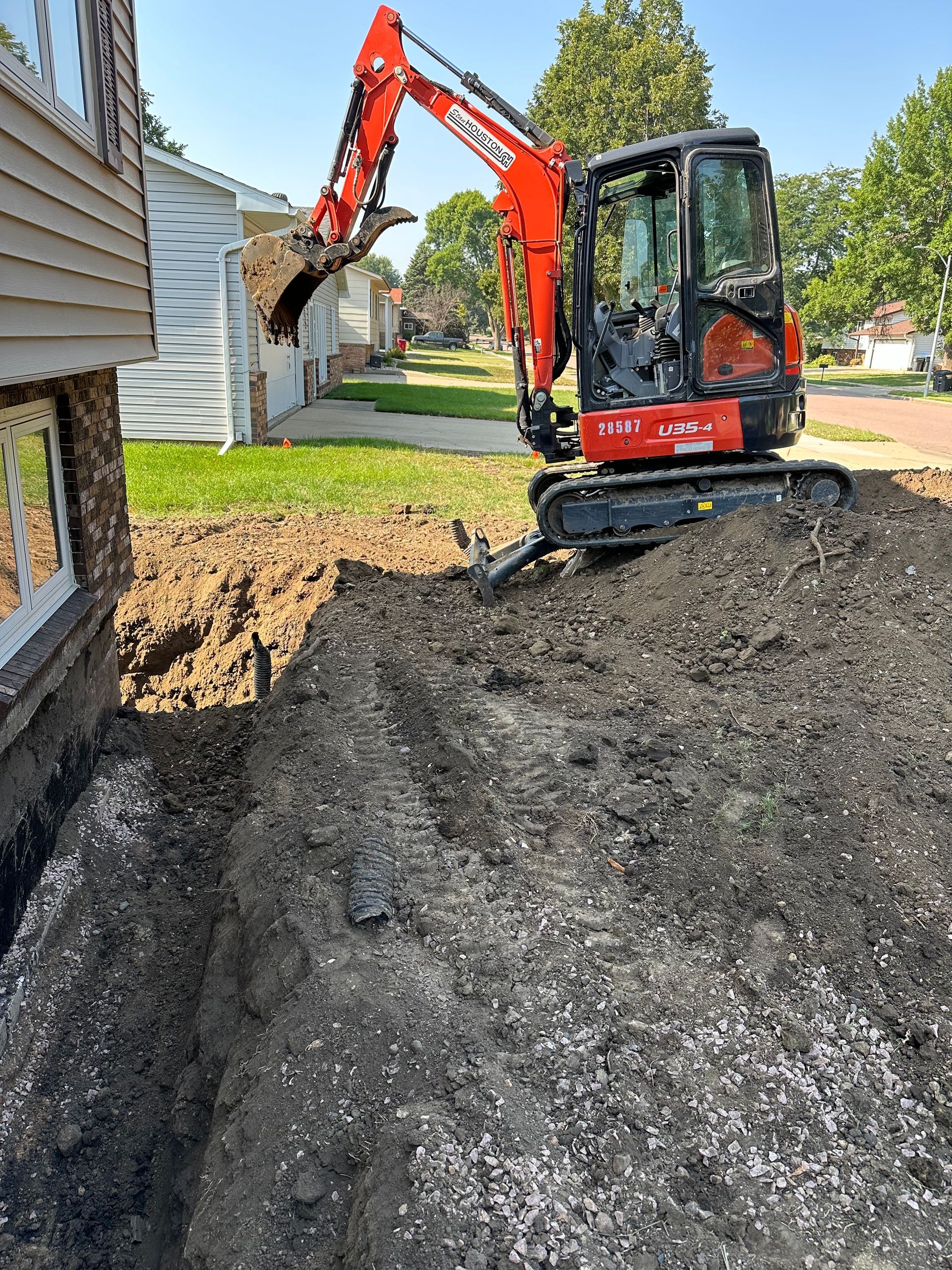 Orange excavator digging trench next to a house on a sunny day.