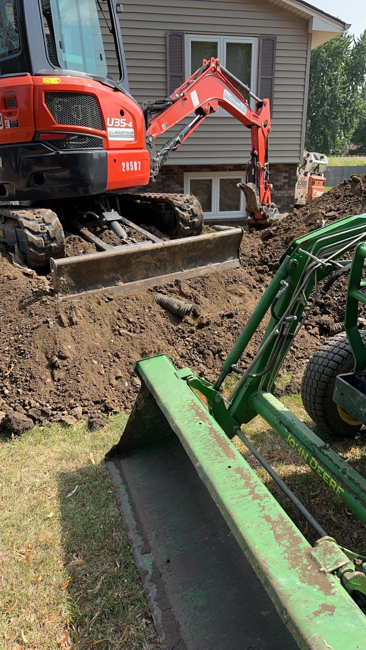 Excavator and tractor digging near a house; dirt piled up with some grass visible.