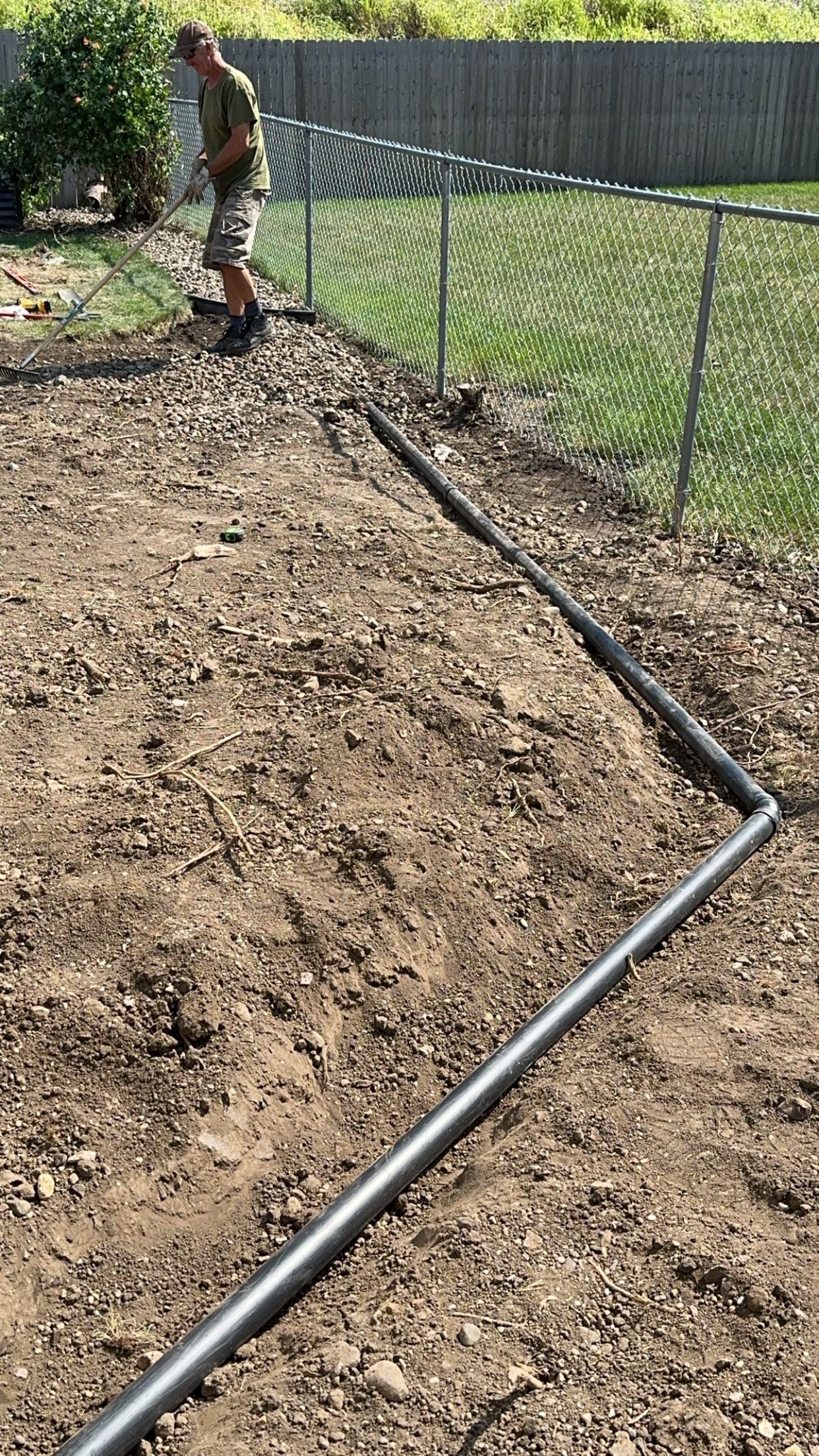 A person installs irrigation pipe near a chain-link fence in a dirt garden bed.
