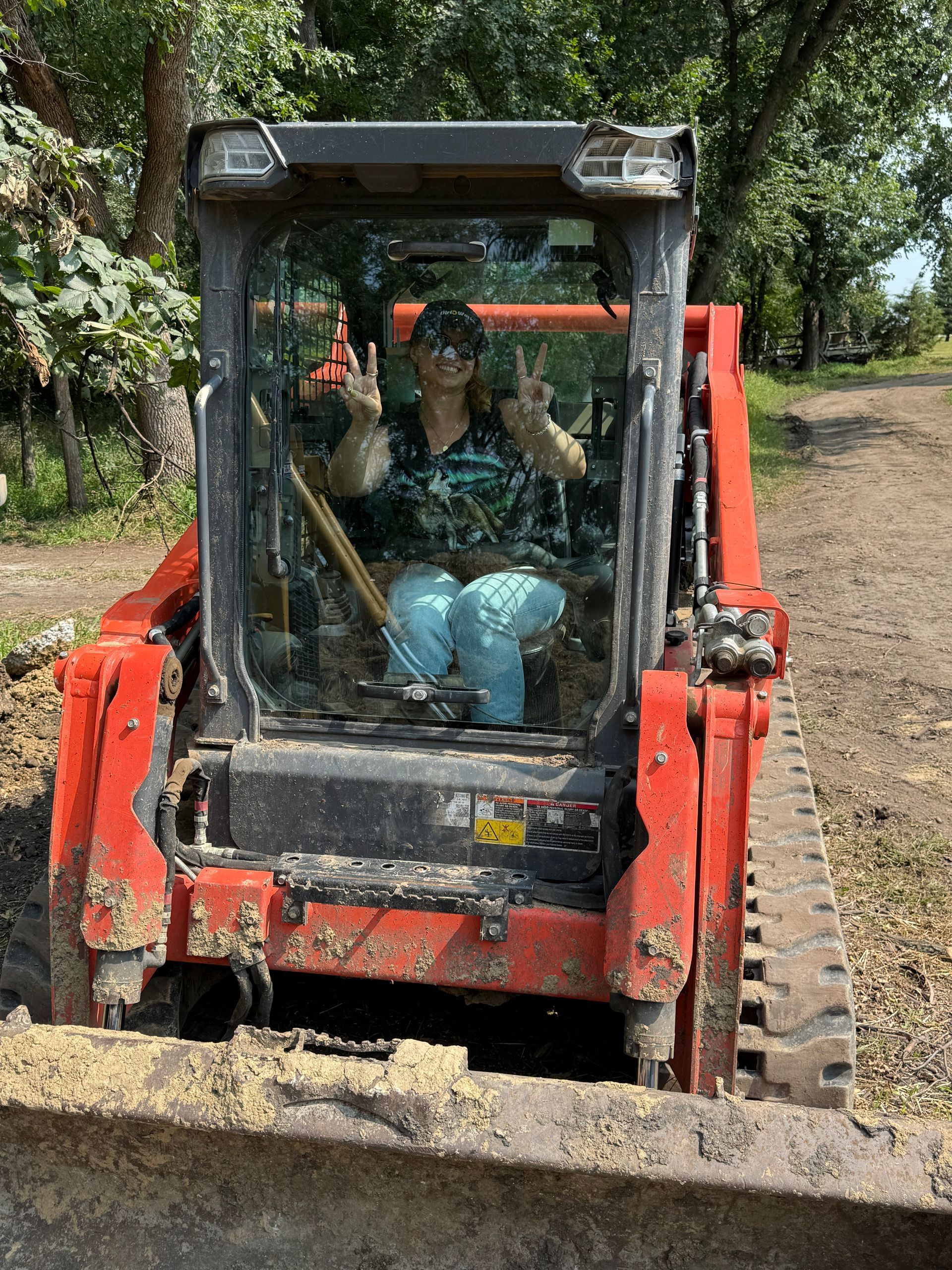 Person in a red skid steer raises two peace signs. The machine is on a dirt path with trees in the background.