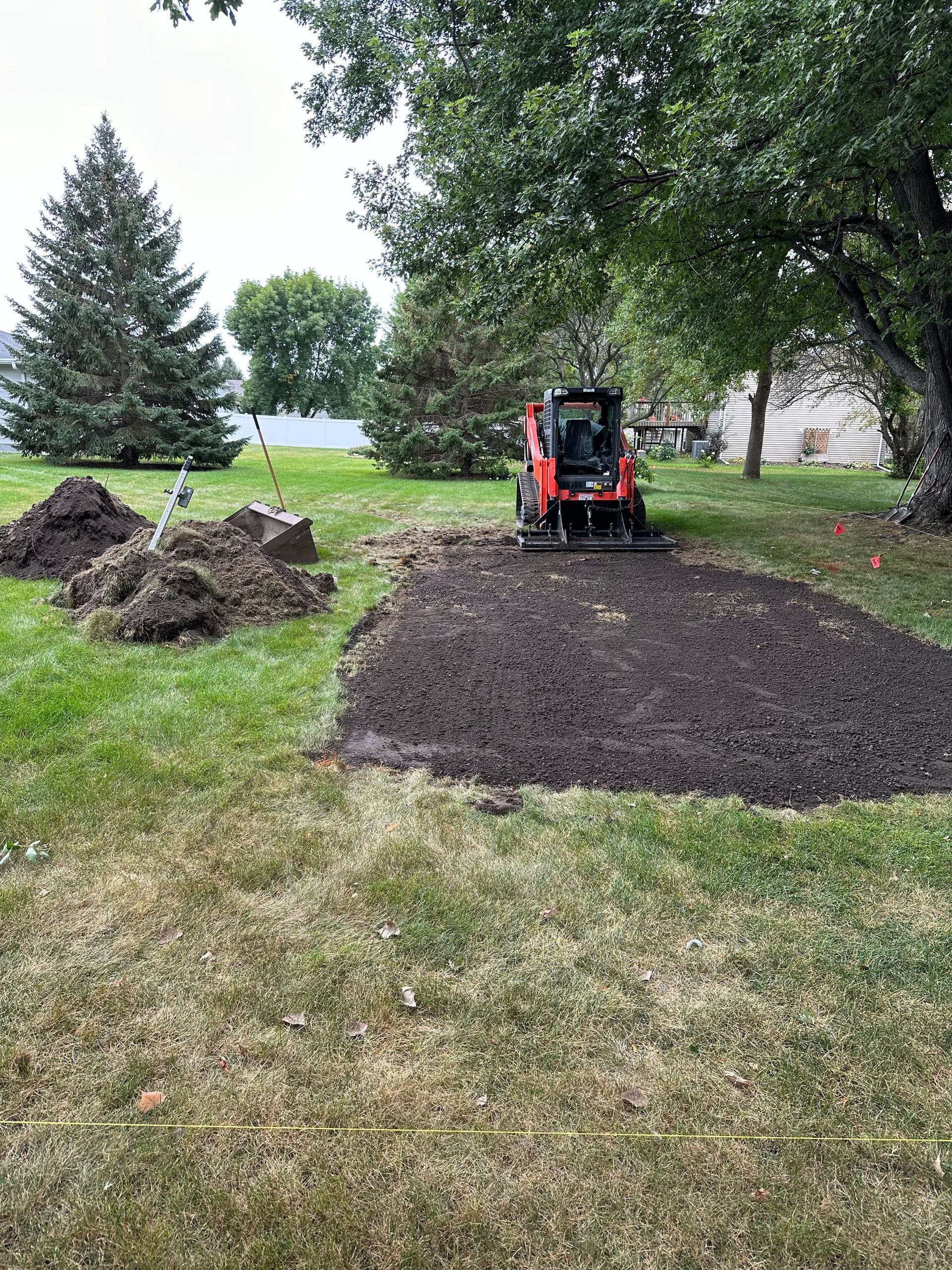 A small orange skid steer levels a patch of dark soil on a lawn, next to a pile of dirt.
