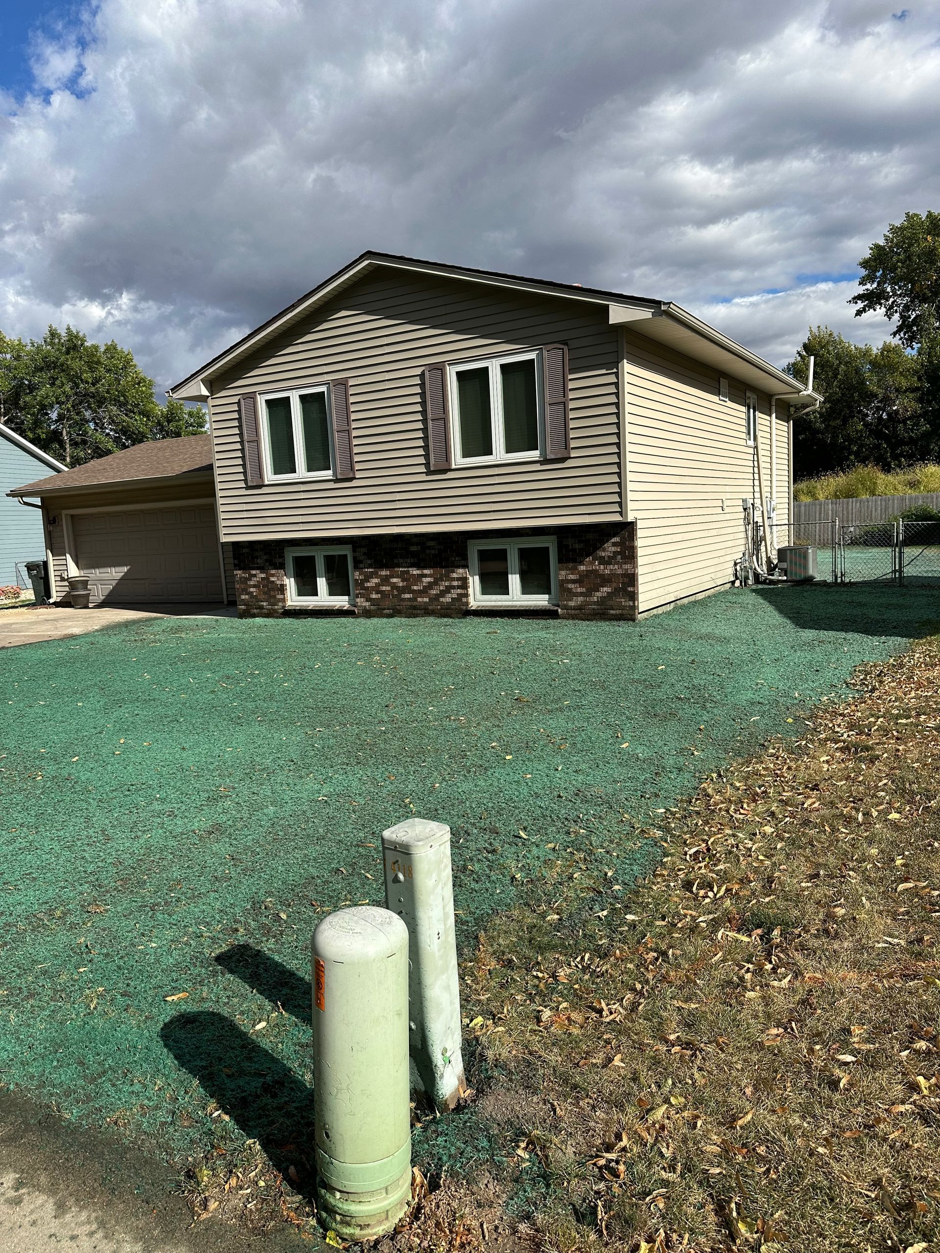 Two-story house with green-tinted grass. Garage on the left, windows, and light siding. Brick at the base. Cloudy sky.