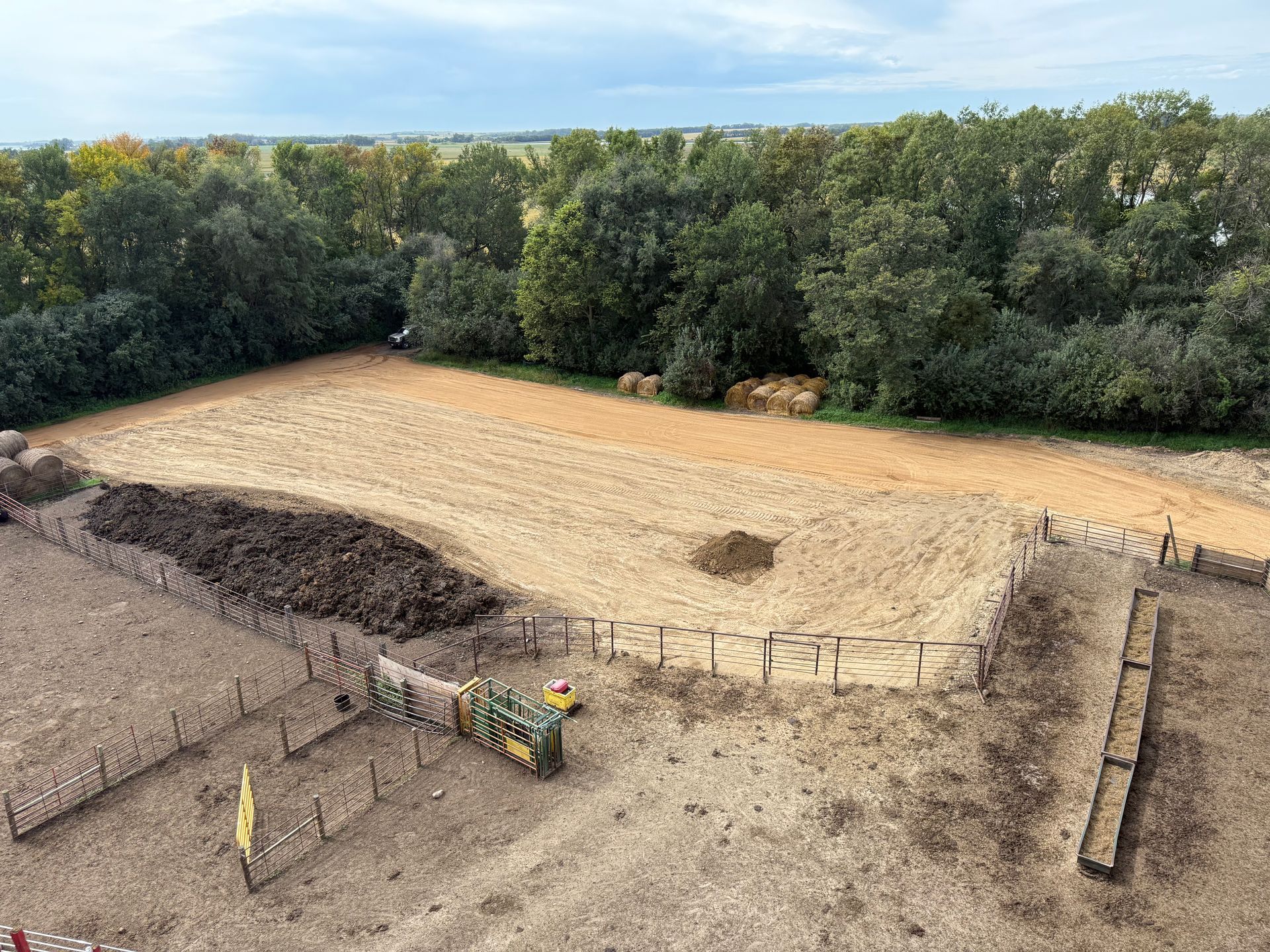 View of a cleared field with piles of dirt, fencing, and trees in the background under a cloudy sky.